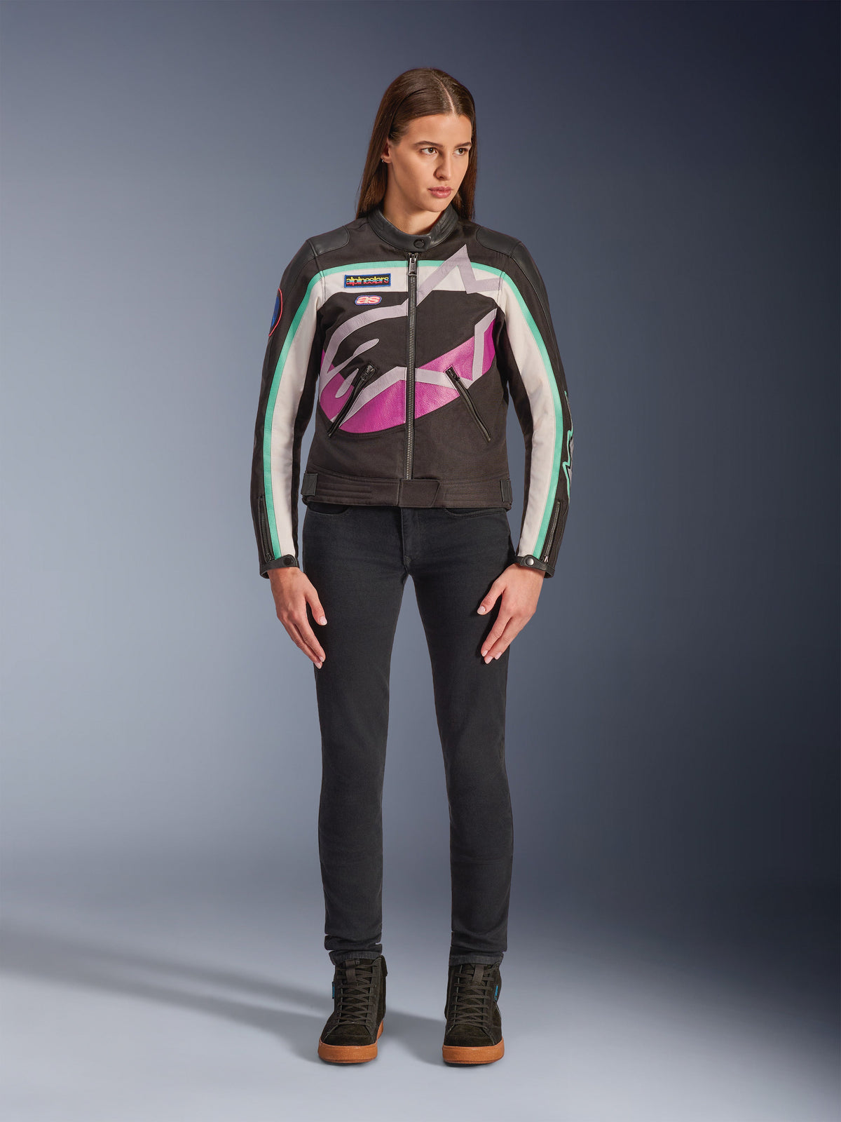 Frau in Alpinestars Idles Damenjacke, Schwarz, Off-White und Lila, von Vintage-Rennsport inspiriertes Design mit großem, umlaufendem Logo und türkisfarbenen Akzenten, kombiniert mit schwarzen Jeans und Motorrad-Sneakern, urbane Motorradbekleidung