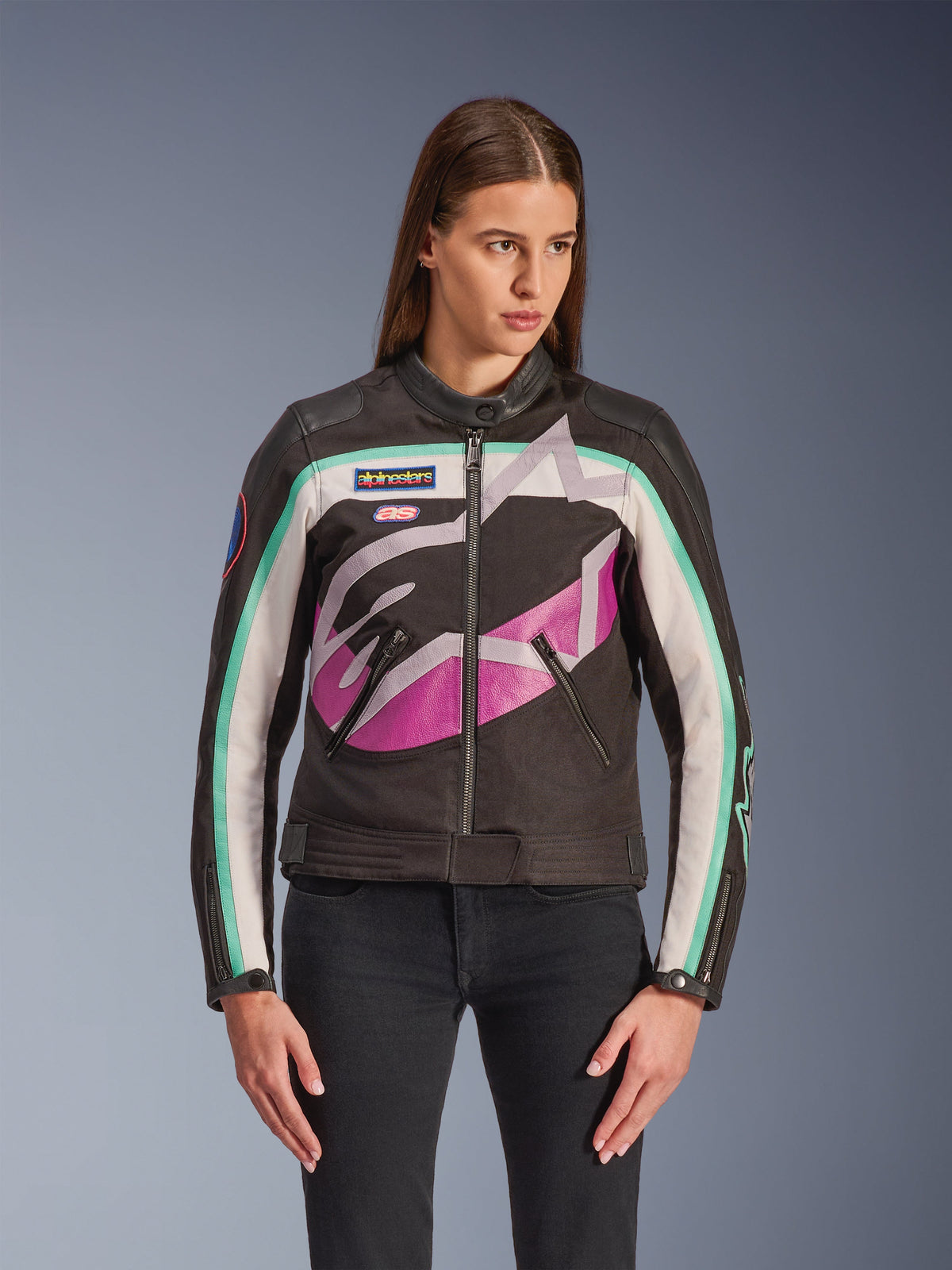 Alpinestars Idles Damenjacke, Motorradjacke, Schwarz mit Color-Block-Design in Off-White, Lila und Türkis, Vintage-Rennsport-inspirierter Stil mit großem abstraktem Logo auf der Brust, Marken-Aufnähern und Frontreißverschluss, getragen von einem Model mit schwarzen Jeans