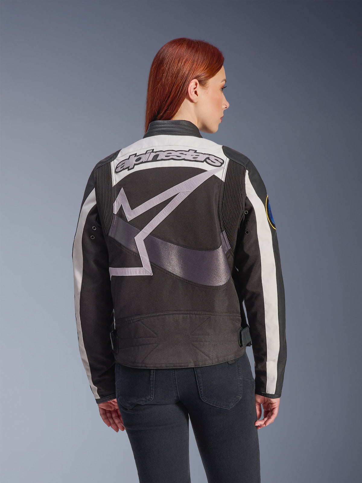 Alpinestars Idles Damenjacke, Schwarz und Off-White, Rückansicht mit großem grauen Stern-Logo und Alpinestars-Schriftzug, getragen von einem rothaarigen Model, vintage-inspirierte urbane Motorradjacke mit Akkordeon-Einsätzen.