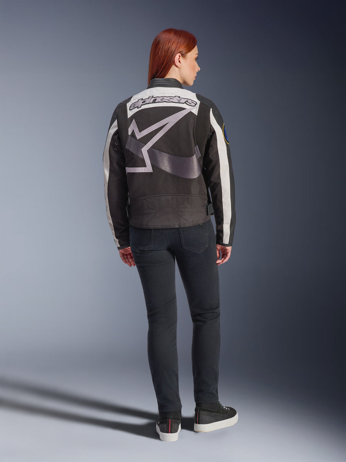 Alpinestars Idles Damenjacke, urbane Motorradjacke, Schwarz und Off-White, Rückansicht mit großem grauem Alpinestars-Logo und Schriftzug, getragen mit schwarzen Slim-Fit-Jeans und Sneakern, Vintage-inspiriertes Design für urbanes Fahren