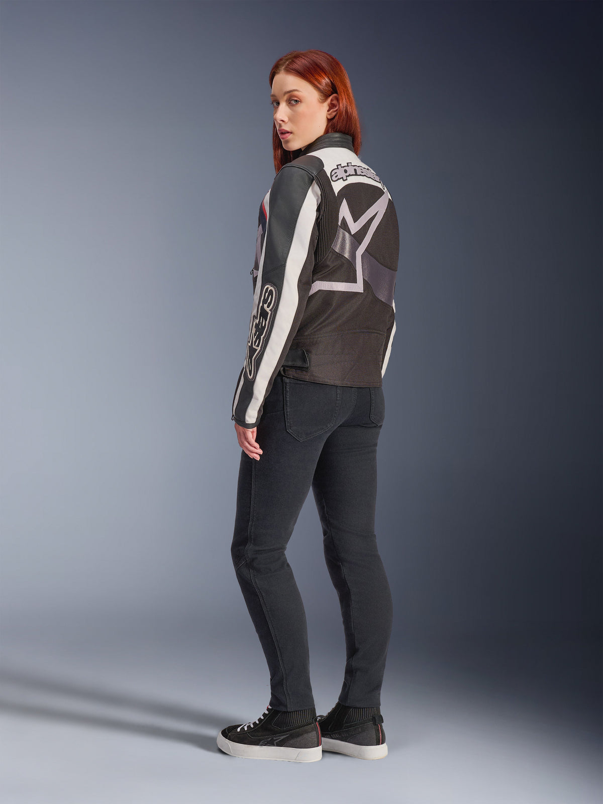 Frau in Alpinestars Idles Jacke, Rückansicht, Off White Flame Red und Schwarz, großes Stern-Logo auf dem Rücken und Alpinestars-Wortmarke auf dem Ärmel, kombiniert mit schwarzer Slim-Fit-Hose und Sneakern, urbane Motorradbekleidung für Damen