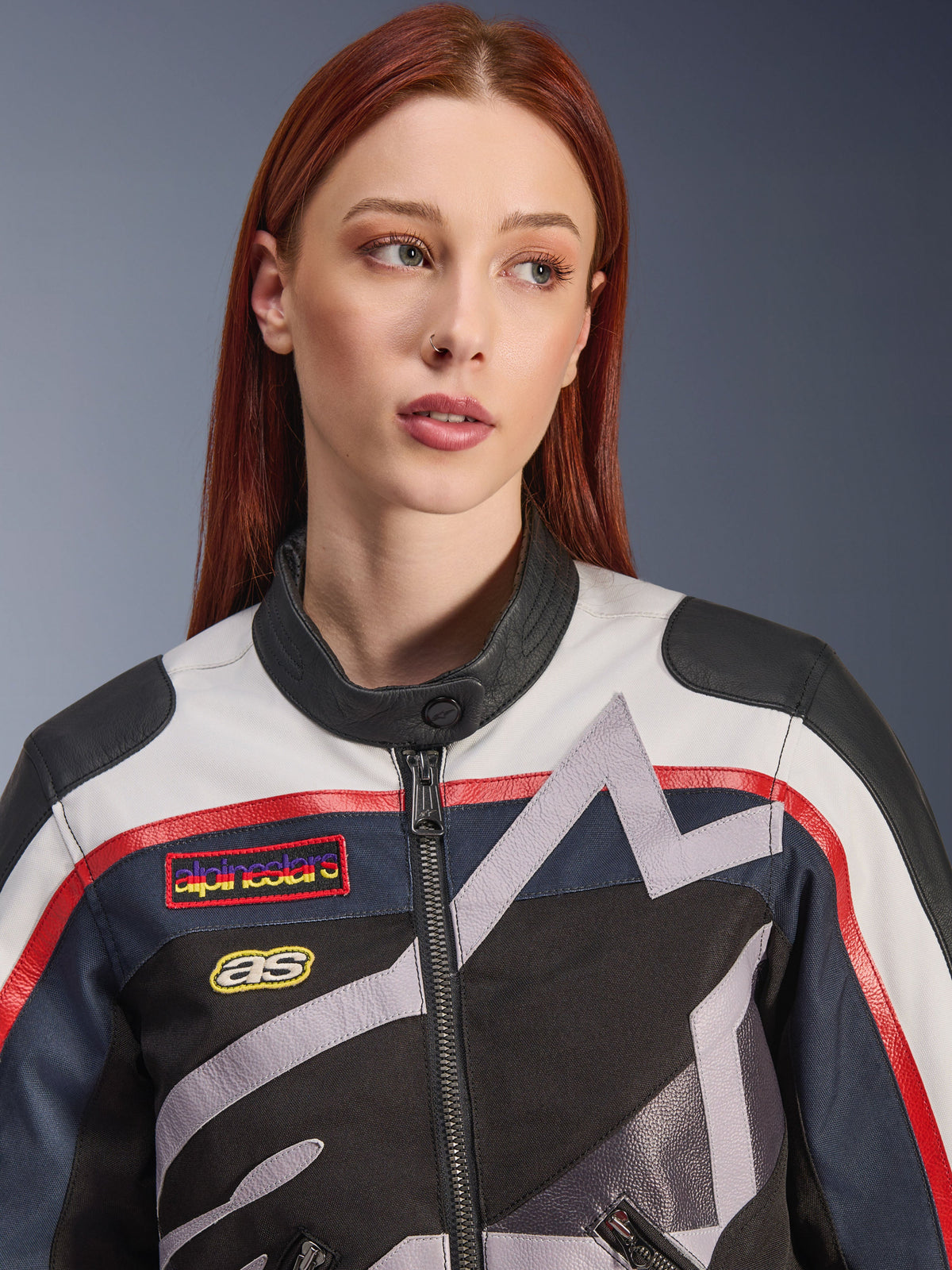 Alpinestars Idles Damenjacke, Nahaufnahme eines weiblichen Models mit roten Haaren, Farbkombination Off White, Flame Red und Schwarz, vintage-inspiriertes Renndesign mit Logo-Aufnähern und grauen geometrischen Sterngrafiken, urbane Motorradbekleidung.