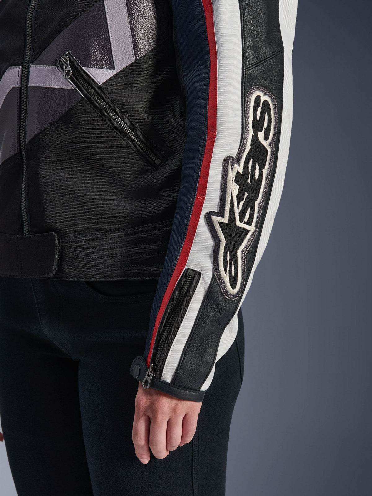 Alpinestars Idles Damenjacke, urbane Motorradjacke, Off-White, Flammenrot, Schwarz, Nahaufnahme des Ärmels mit einem großen bestickten Alpinestars-Logo, rote und weiße Rennstreifen, Konstruktion aus schwarzem Textil und Leder sowie Akzente durch Metall-Reißverschlüsse.