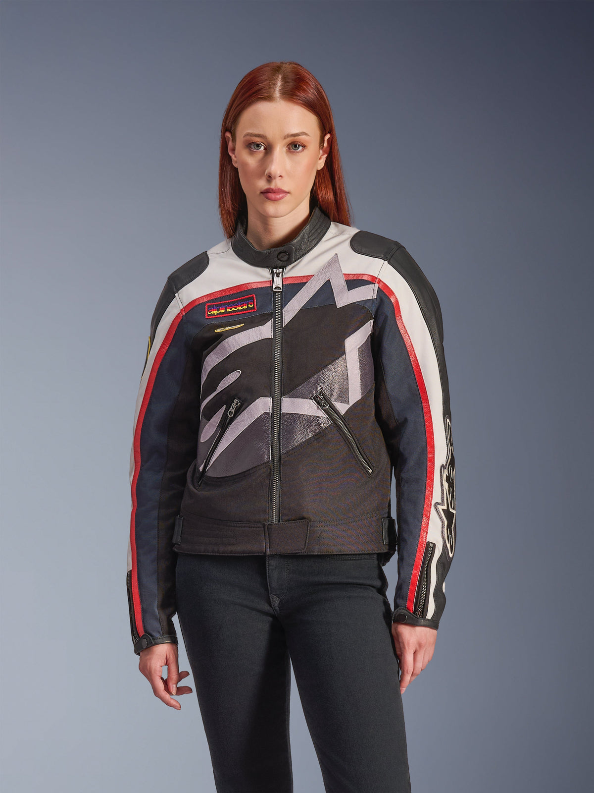 Alpinestars Idles Jacke, Damen-Motorradjacke, Off-White, Flame-Red und Schwarz, mit großem stilisiertem Stern-Logo, Vintage-inspirierten Brust-Aufnähern, hohem Biker-Kragen, Reißverschlusstaschen und gestreiften Ärmeln, getragen von einem weiblichen Model mit dunkler Jeans.