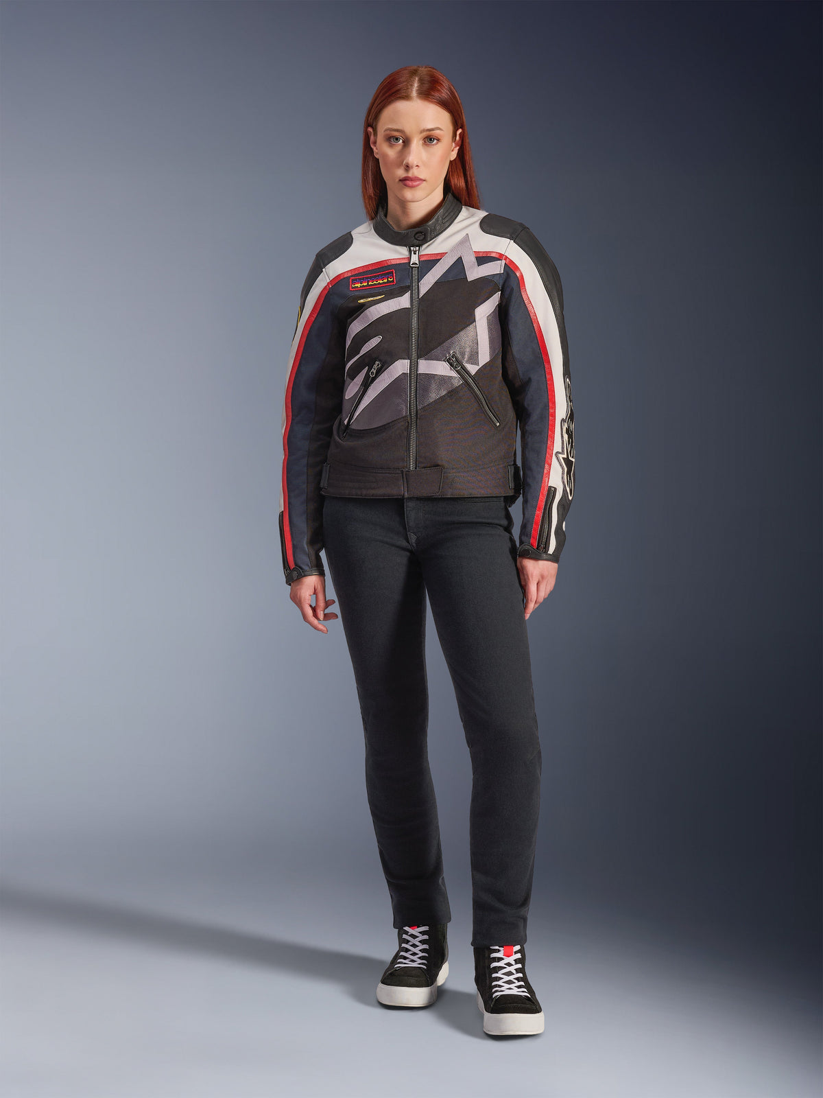 Weibliches Modell in der Alpinestars Idles Jacke, Motorradjacke für Damen, Off-White, Flame Red und Schwarz, mit einem großen, umlaufenden Stern-Logo und Vintage-inspiriertem Branding, kombiniert mit einer dunklen Slim-Fit-Fahrhose und schwarzen High-Top-Sneakern, urbane zeitgenössische Motorradausrüstung