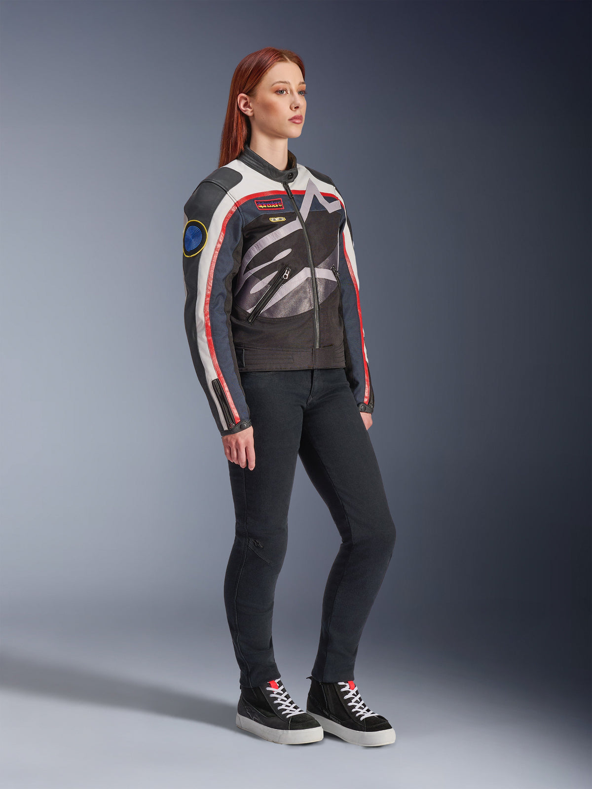 Alpinestars Idles Damenjacke, Motorradjacke, Off-White, Flame Red und Schwarz, mit einem großen, umlaufenden Alpinestars-Logo und von Vintage-Rennsport inspirierten Aufnähern, kombiniert mit schwarzer Hose und High-Top-Sneakern, moderne urbane Fahrausrüstung