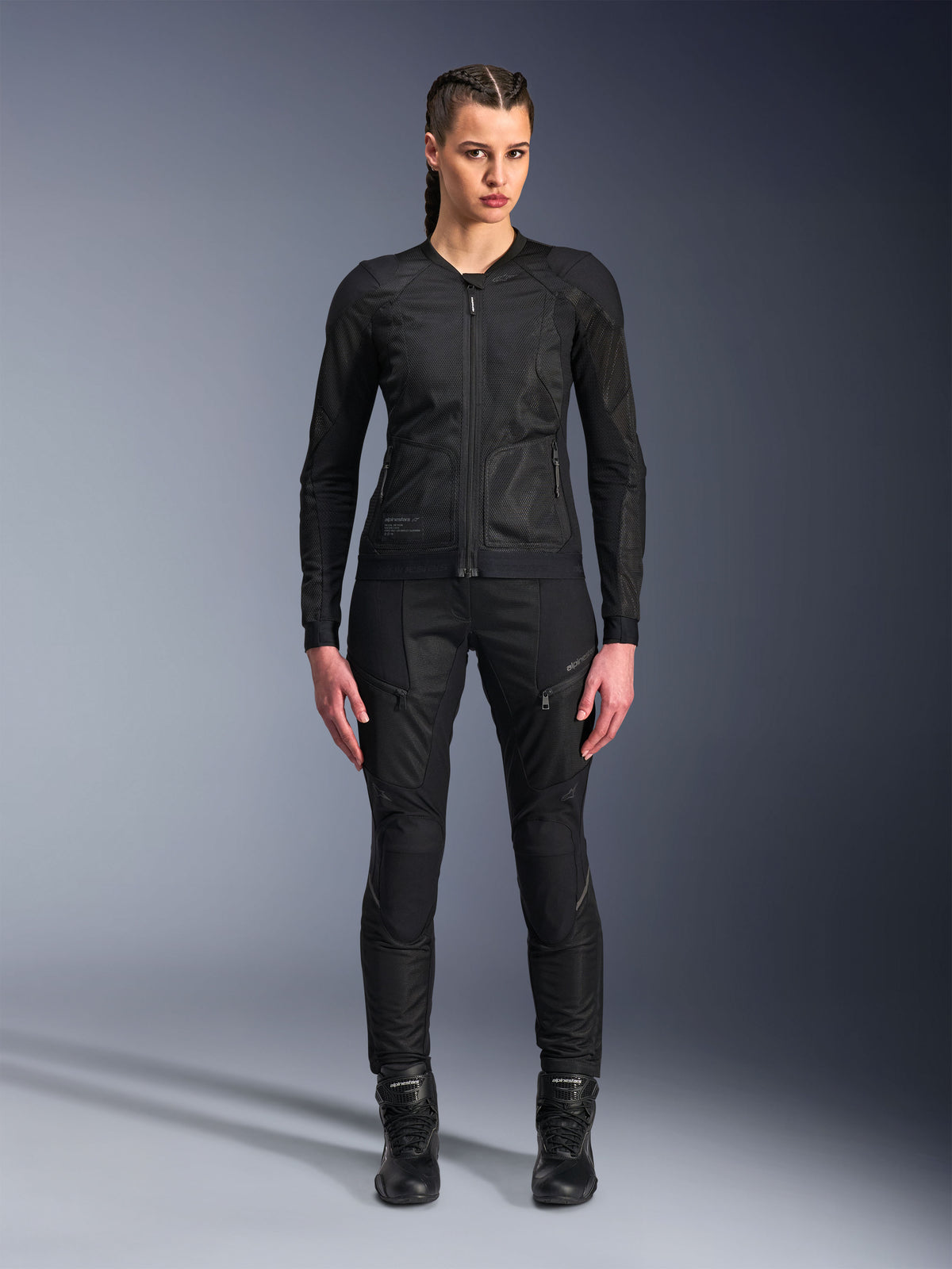 Veste Alpinestars Stella Troop-Air, veste de moto pour femme, noire, construction technique en maille 2D avec un design minimaliste, portée avec un pantalon de moto et des bottes noirs, style de moto urbain contemporain.