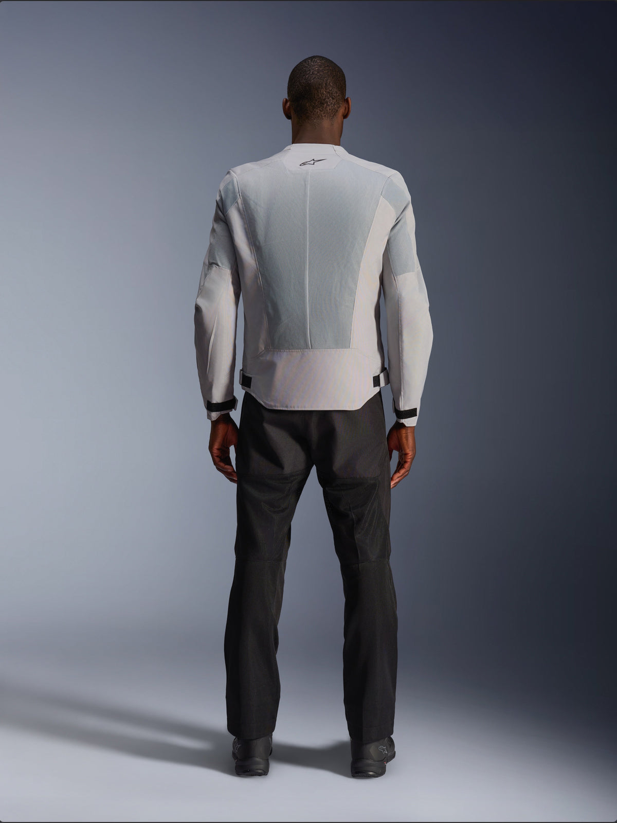 Alpinestars C-1 Air Jacket in Silver, viewed from the back on a male model, textile motorcycle jacket featuring large tech mesh ventilation panels, small black logo at the neck, and waist adjusters, worn with black riding pants for an urban contemporary look.