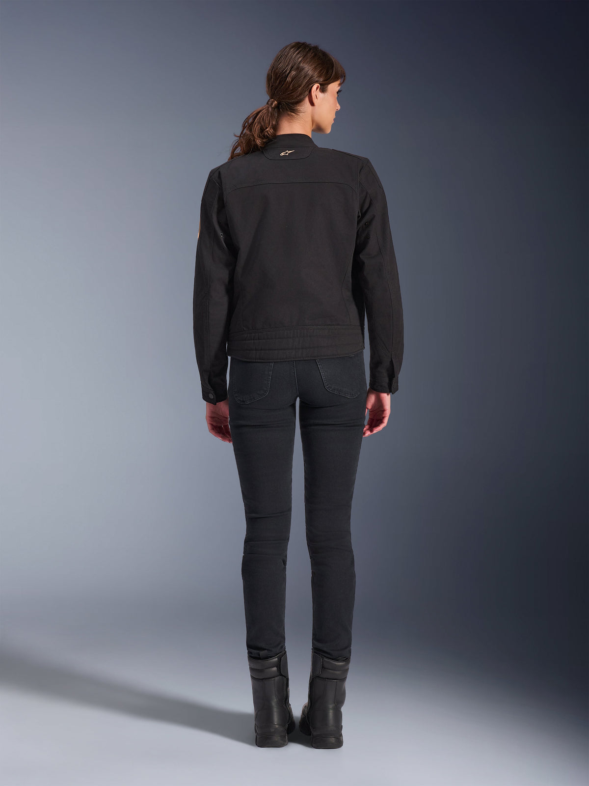 Veste Stella Grace Alpinestars, veste de moto urbaine pour femme, noire, vue de dos montrant le design d'inspiration workwear, portée avec un pantalon slim noir et des bottes noires, photo en studio