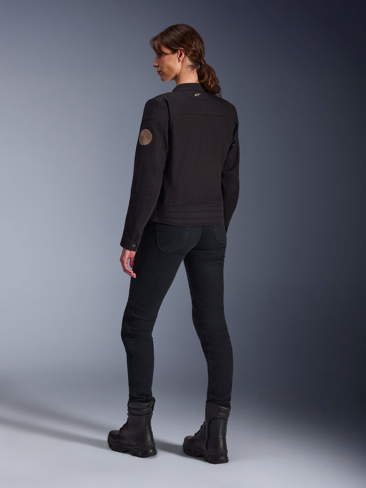 Mannequin portant la veste Alpinestars Stella Grace en noir, vue de dos mettant en évidence l'écusson Oscar vintage en spirale sur la manche et le petit logo étoile sur le col, assortie d'un pantalon de moto noir et de bottes noires pour la conduite urbaine