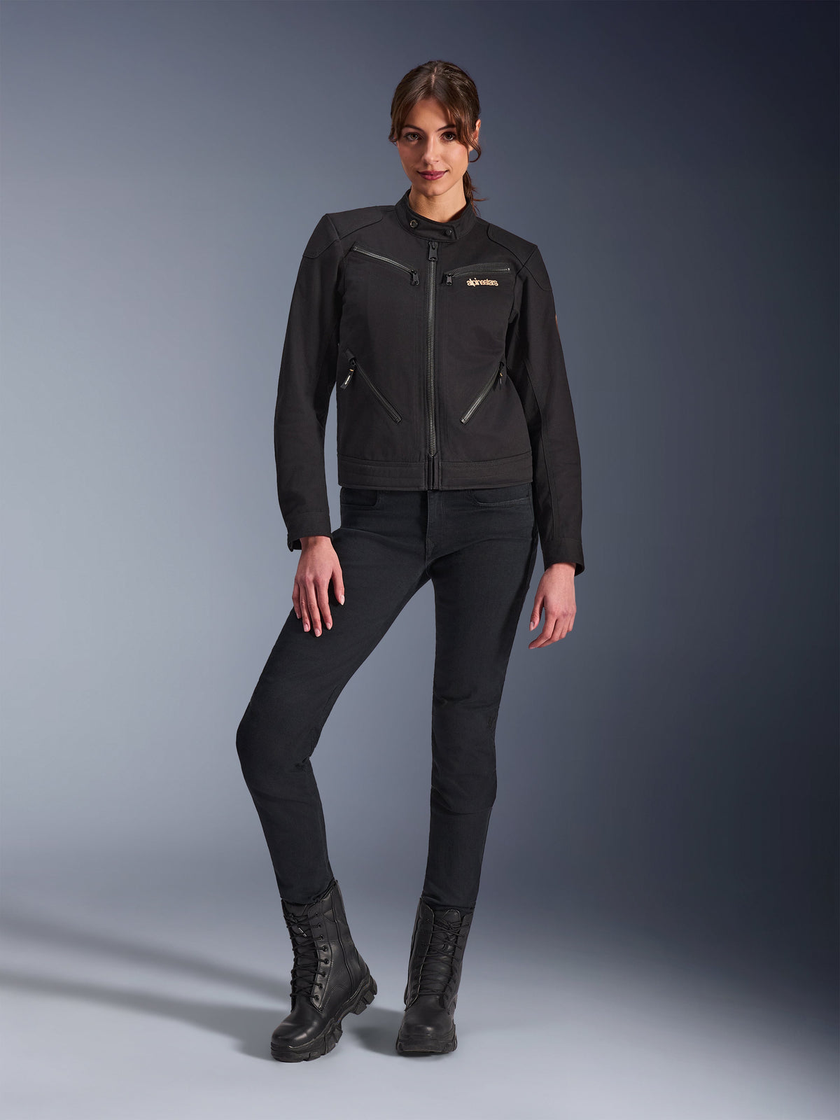 Femme portant la veste Alpinestars Stella Grace en noir, veste de moto urbaine à l'esthétique workwear, dotée de poches avant zippées et d'un logo sur la poitrine, associée à un pantalon noir ajusté et des bottes noires, équipement de moto contemporain pour femme