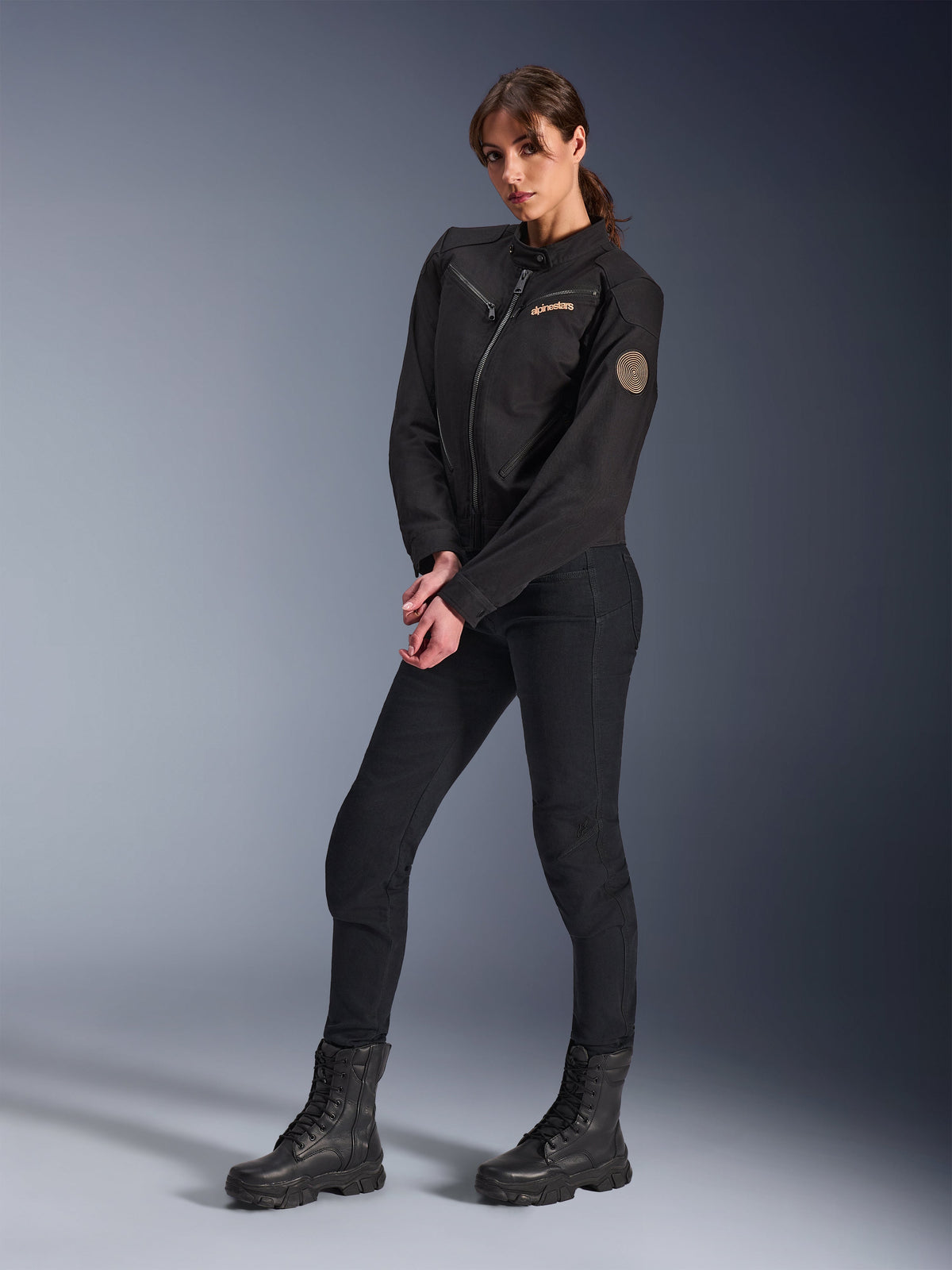 Mannequin femme portant la veste Alpinestars Stella Grace, veste de moto urbaine noire, assortie d'un pantalon de moto et de bottes noirs, design d'inspiration workwear avec un logo brodé bronze sur la poitrine et un écusson circulaire en spirale sur la manche