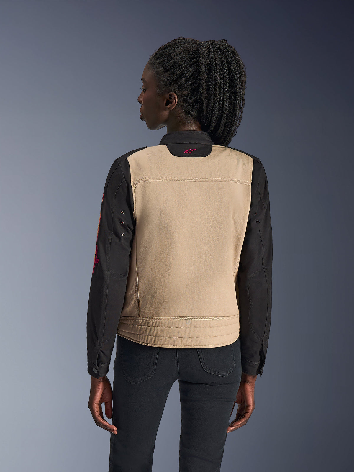Veste Alpinestars Stella Grace pour femme, veste de moto urbaine, coloris Aluminium et Noir, vue de dos montrant un empiècement dorsal beige et des manches noires, design inspiré du style workwear avec un petit logo rose sur le col, associée à un pantalon foncé