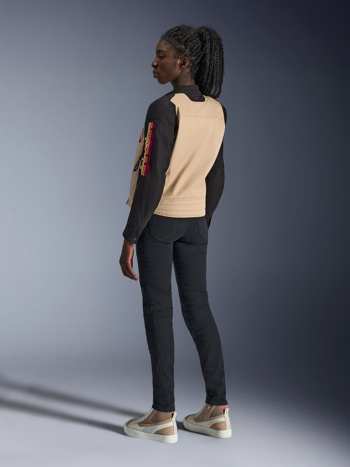 Veste Alpinestars Stella Grace pour femme, veste de moto urbaine, coloris Aluminum Black avec empiècement arrière beige et manches noires, vue de dos de profil sur un mannequin femme, avec logo coloré brodé sur la manche et épaules plissées.