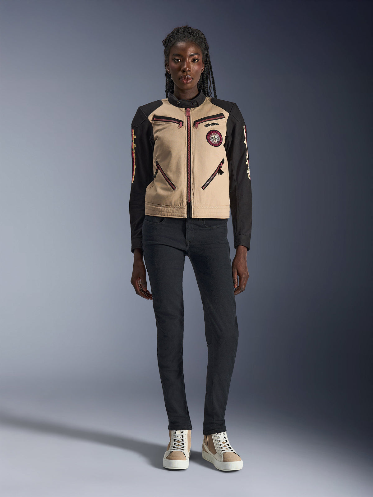 Veste Alpinestars Stella Grace pour femme, veste de moto urbaine, construction textile beige et noire, avec fermetures éclair à l'avant et patch Oscar en spirale brodé, portée avec un pantalon noir et des baskets montantes, équipement de moto d'inspiration workwear contemporaine