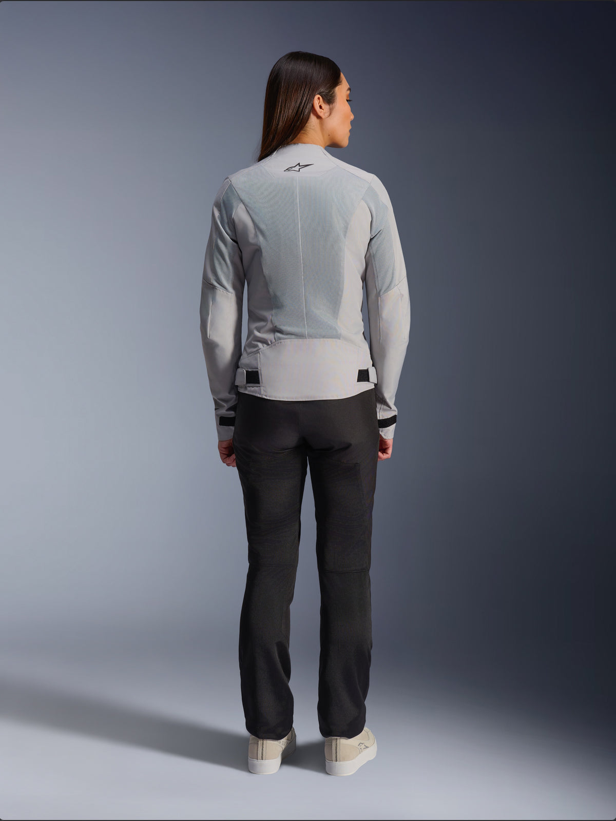 Woman wearing Alpinestars Stella C-1 Air Jacket in Silver, back view, featuring large tech mesh panels for ventilation and a reflective star logo, paired with black riding pants and white sneakers, urban contemporary motorcycle gear