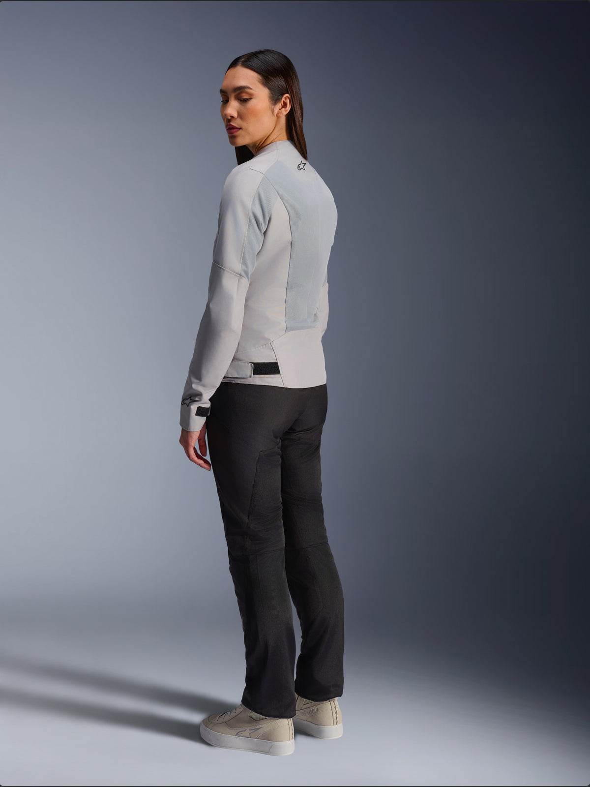Woman wearing Alpinestars Stella C-1 Air Jacket in Silver, back and side view showing tech mesh panels and reflective shoulder logo, paired with black motorcycle pants and beige sneakers, contemporary urban riding aesthetic