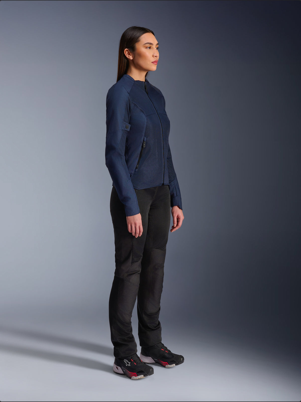 Frau in der Alpinestars Stella C-1 Air Jacke in Blau, urbane Motorradjacke mit leichten Mesh-Einsätzen zur Belüftung, kombiniert mit schwarzer Motorradhose und technischen Schuhen, schlankes Profil und dezentes Branding, Studio-Seitenansicht