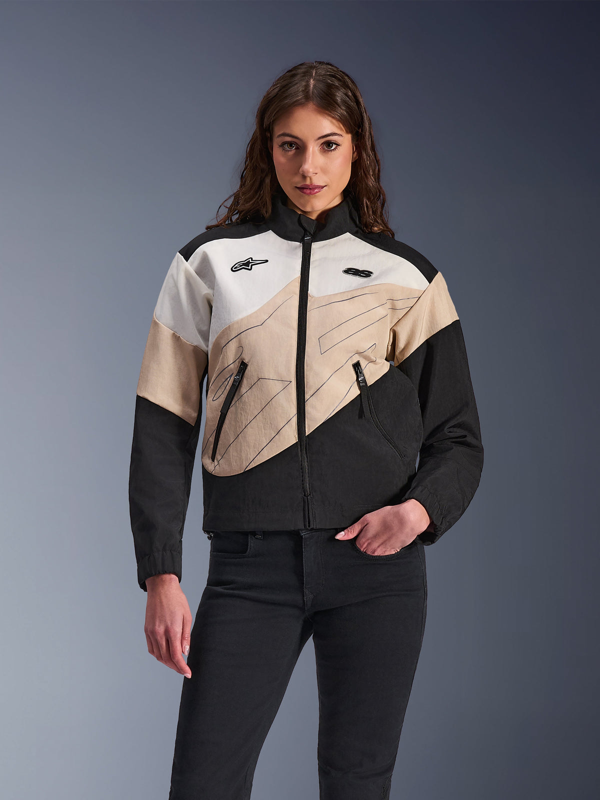 Alpinestars Stella Moshell Jacket, urban motorjas voor dames, asymmetrisch kleurgeblokt ontwerp in zwart, zand en off-white, voorzien van ritssluiting aan de voorzijde en steekzakken met rits, gedragen met een zwarte broek voor een eigentijdse streetwear-look.