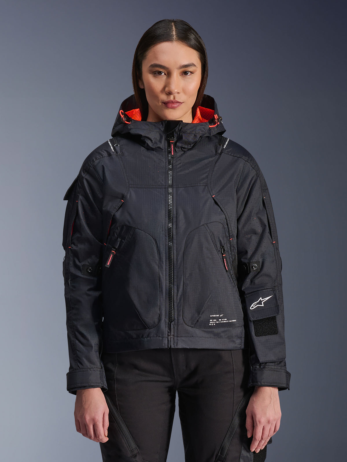 Alpinestars Molly Air Jacke, Damen-Motorradjacke im urbanen Stil, Schwarz mit Innenfutter in Fluo Coral, getragen von einem weiblichen Model, Kapuzendesign mit technischer Textilkonstruktion, Reißverschlusstaschen und Alpinestars-Logo am Ärmel