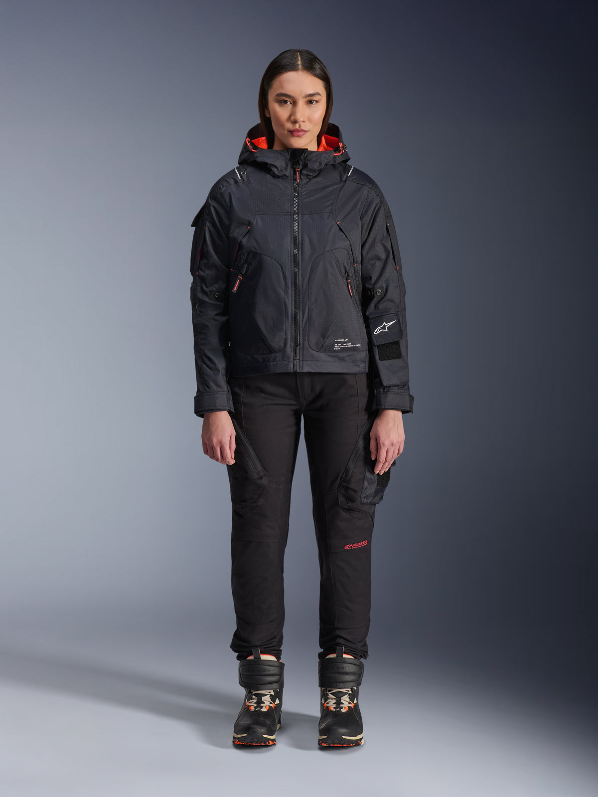 Weibliches Model in der Alpinestars Molly Air Jacke in Schwarz und Fluo Coral, Utility-inspiriertes Design mit Kapuze und korallenfarbenem Innenfutter, kombiniert mit schwarzer Motorradhose und Stiefeln, urbane zeitgenössische Motorradausrüstung