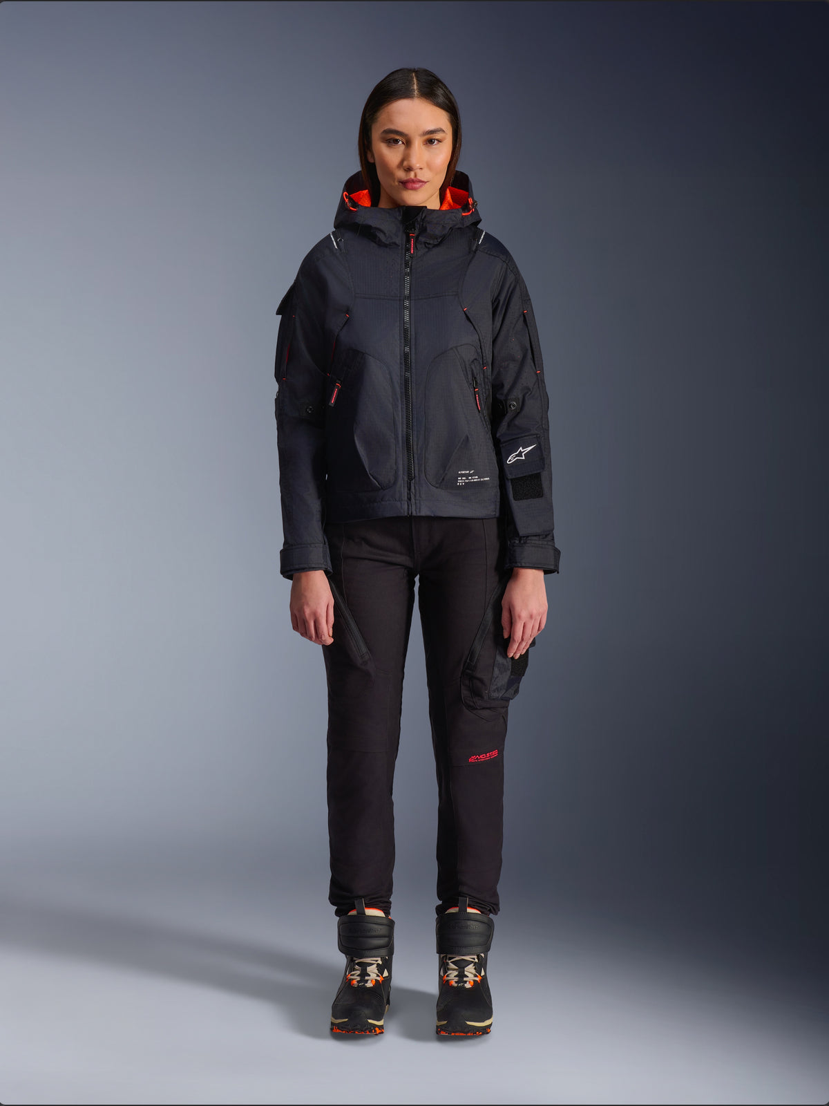Alpinestars Actea damesbroek, zwarte motorbroek met rode tekstdetails en cargozakken, gedragen door een vrouwelijke motorrijder met een zwart jack met capuchon, moderne urban motorkleding