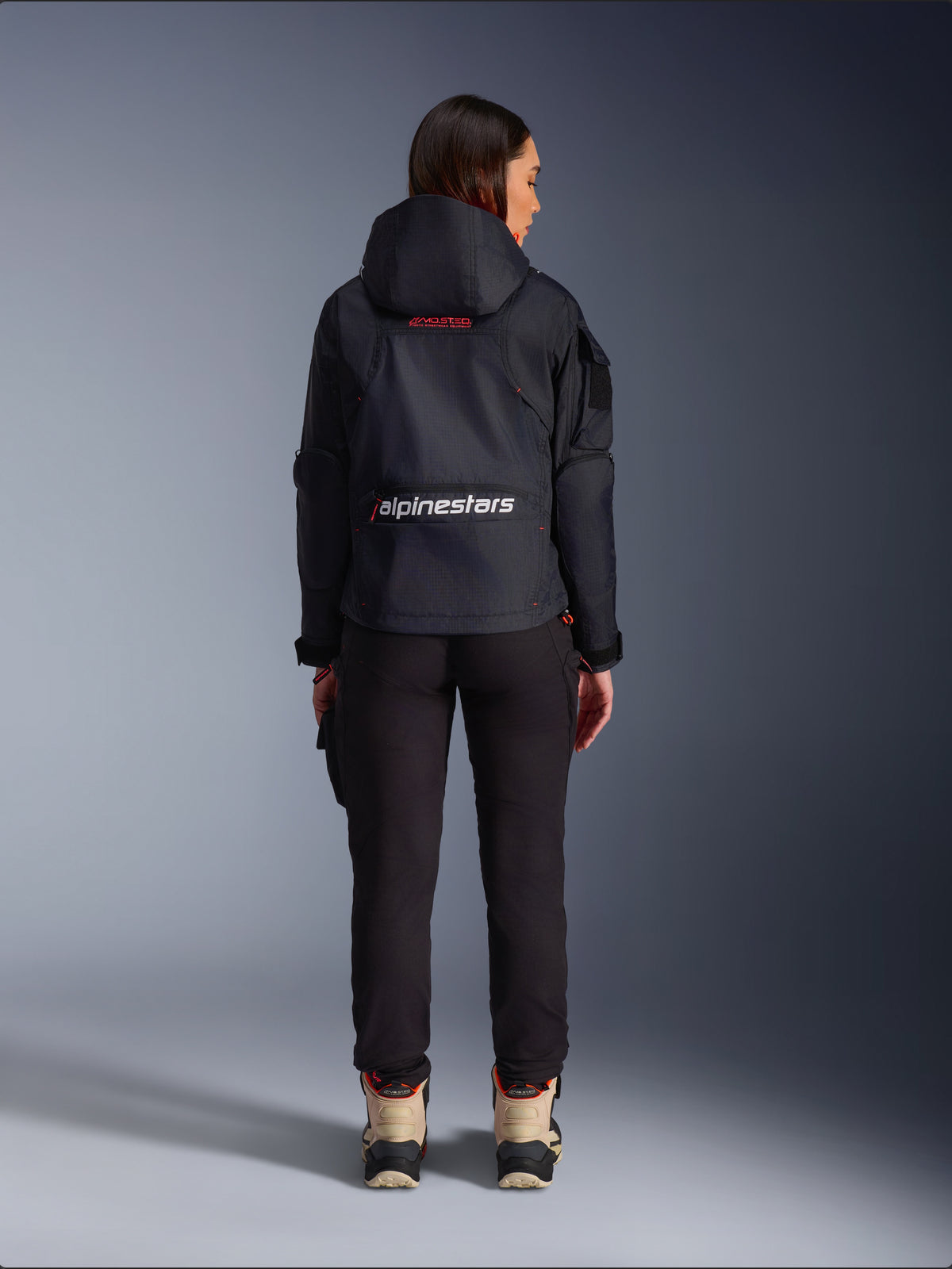 Alpinestars Molly Air Jacke, Urbane Motorradjacke, Schwarz und Fluo Coral, Rückansicht mit Kapuze und großem weißen Branding auf dem unteren Rücken, getragen mit schwarzer Hose und beigefarbenen Motorradstiefeln, leichte Textilkonstruktion