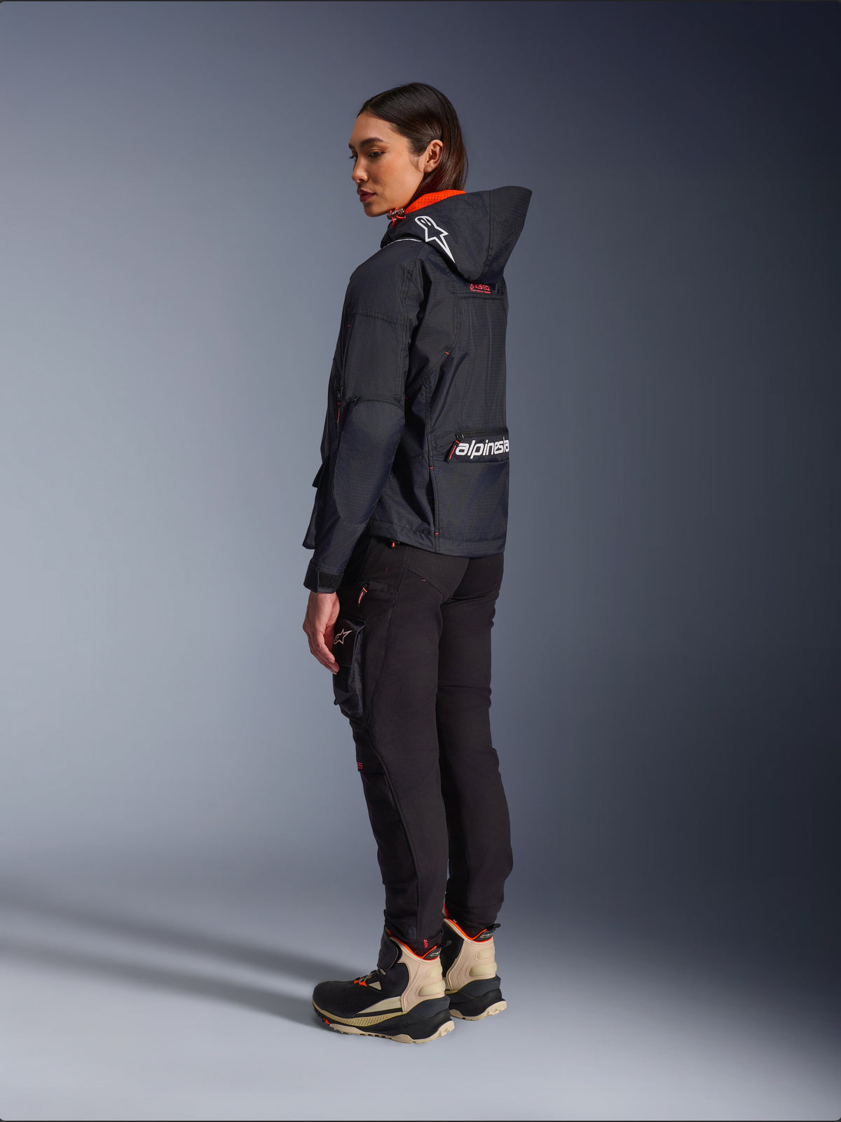 Motorradfahrerin in der Alpinestars Molly Air Jacke in Black Fluo Coral, Seitenansicht von hinten zeigt die Kapuze mit Astars-Logo und großem Alpinestars-Branding auf dem Rücken, kombiniert mit schwarzer Cargo-Motorradhose und sandfarbenen sowie schwarzen Adventure-Stiefeln, moderne urbane Motorradausrüstung für Damen