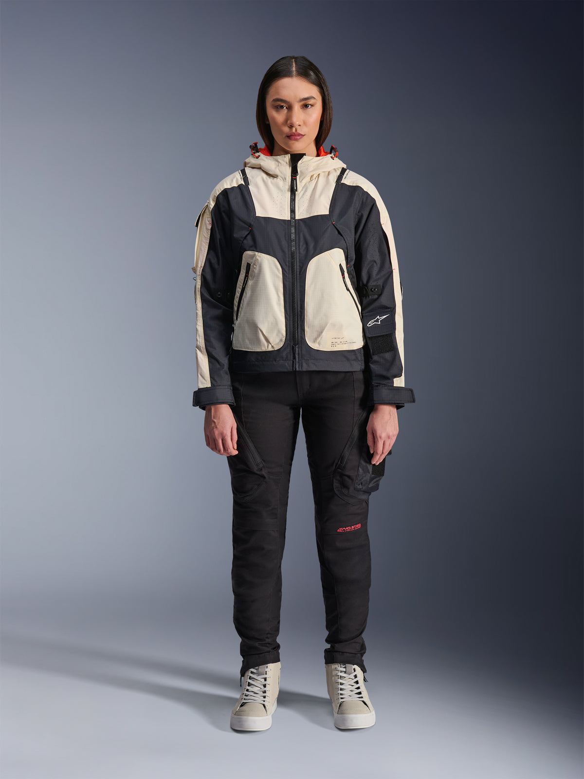 Female model wearing the Alpinestars Molly Air Jacket in Black Almond, featuring a color-blocked design with cream and black panels and a red-lined hood, paired with black motorcycle pants and cream sneakers, studio shot of urban contemporary riding gear.