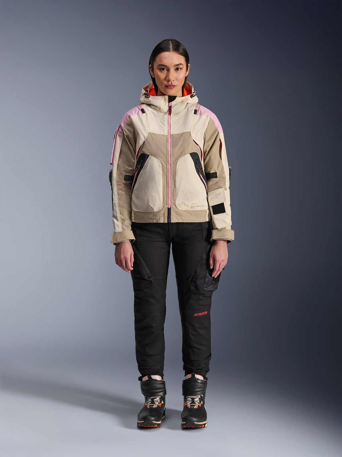 Female model wearing the Alpinestars Molly Air Jacket in Sand Pink Almond, paired with black technical riding pants and boots, front full-body view, urban motorcycle jacket with utility-inspired design and pink accents