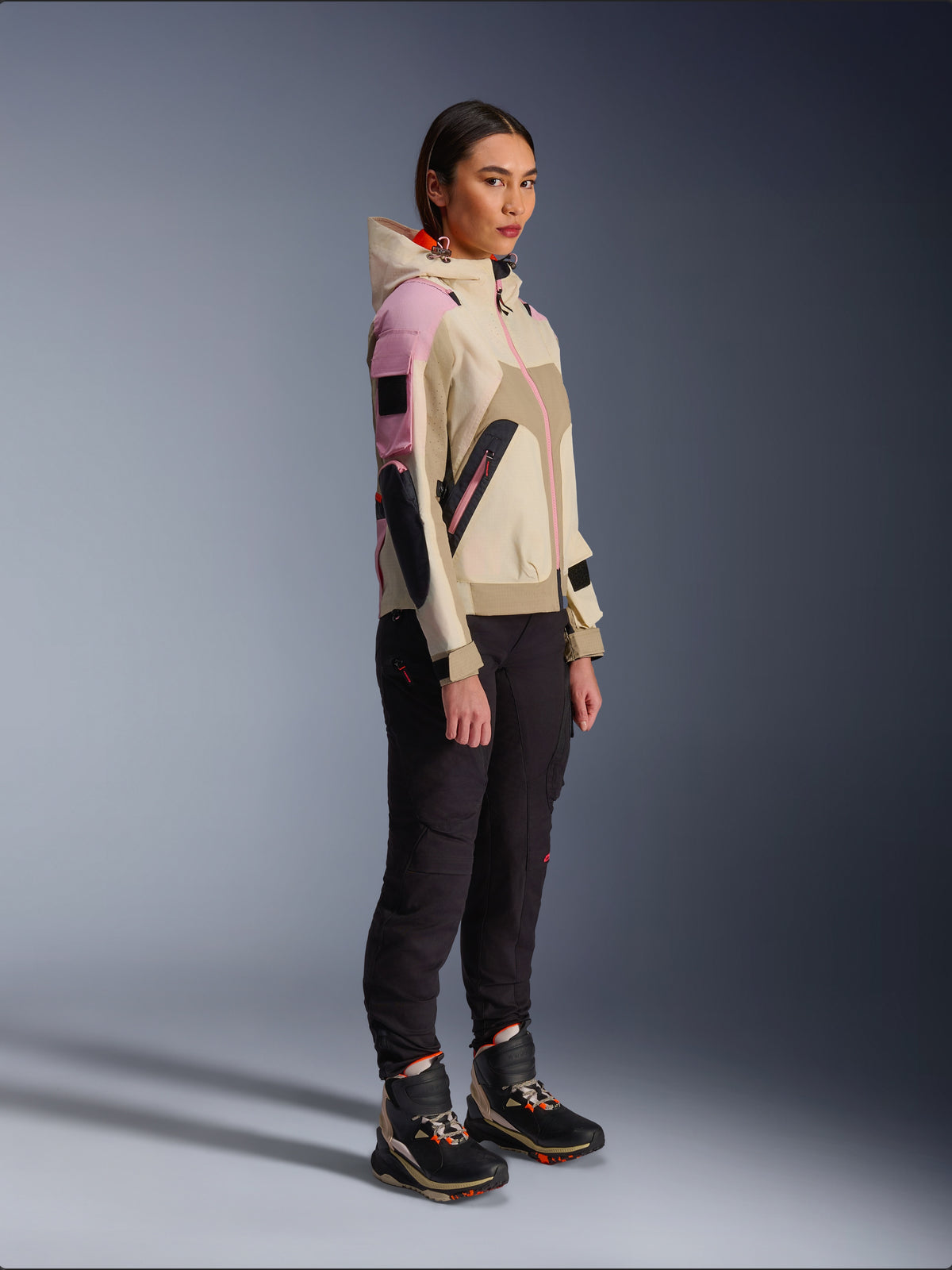 Female model wearing the Alpinestars Molly Air Jacket in Sand Pink Almond, paired with dark technical pants and high-top riding boots, urban motorcycle jacket featuring a hood and multi-color paneling, contemporary streetwear-inspired design