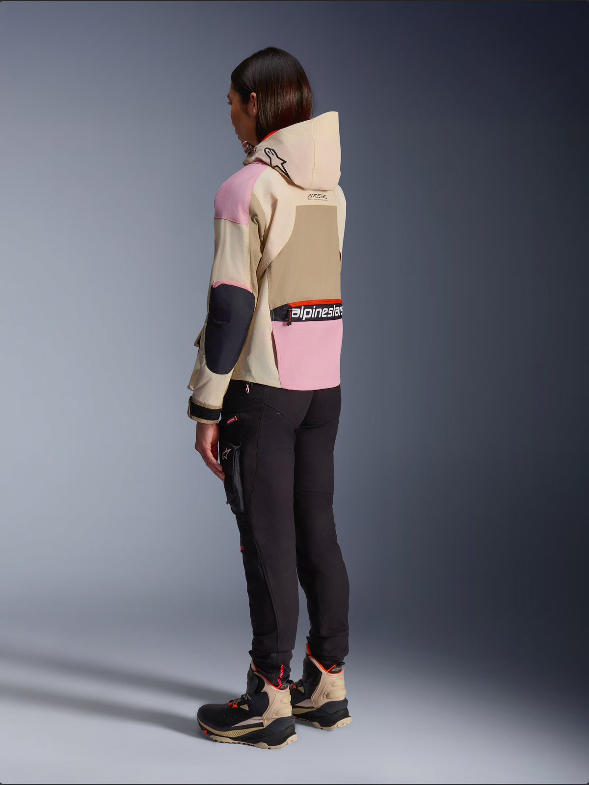 Female rider wearing the Alpinestars Molly Air Jacket in Sand Pink Almond, featuring a multi-tonal beige and pink design, shown from a rear side view with black riding pants and matching technical boots, urban motorcycle style with a hood.