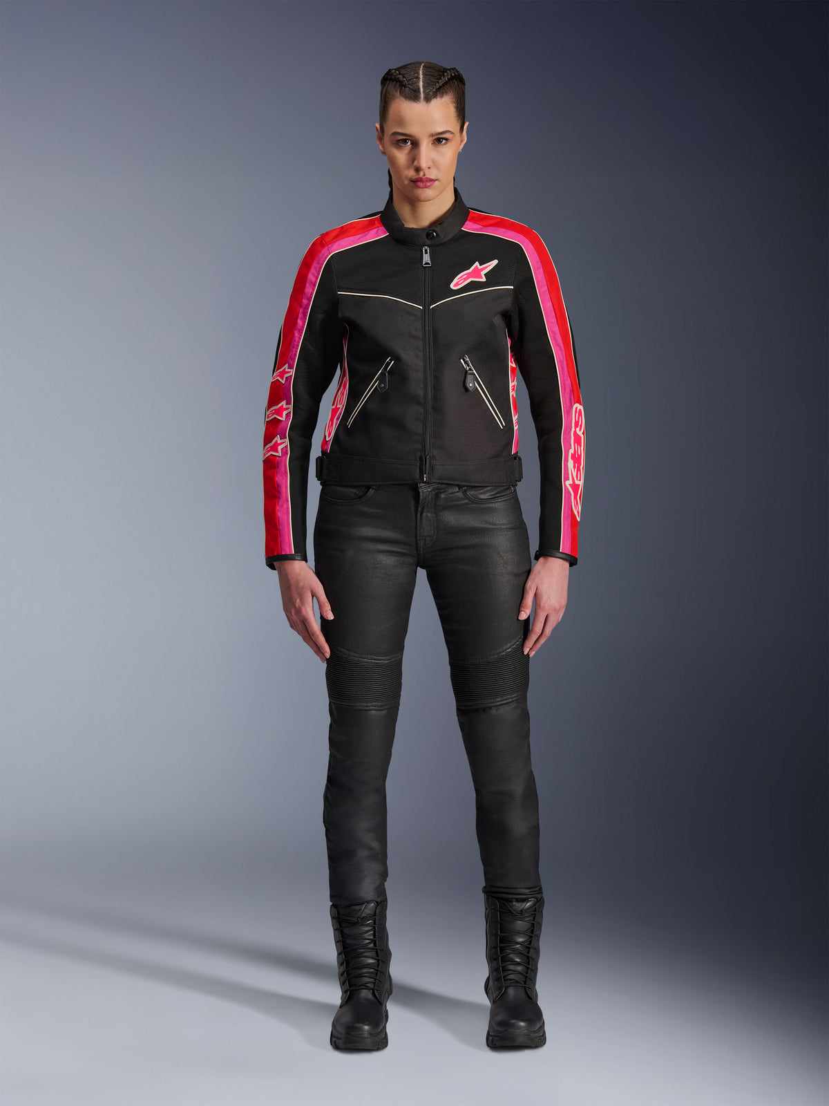 Motorradfahrerin in der Alpinestars Stella T-Dyno Air Jacke, Schwarz mit hellroten und fuchsiafarbenen Akzenten, kombiniert mit schwarzer Motorradhose und Schnürstiefeln, mit 3D-Mesh-Einsätzen, taillierter Passform und pinkfarbenen Stern-Logos, urbanes Motorrad-Outfit
