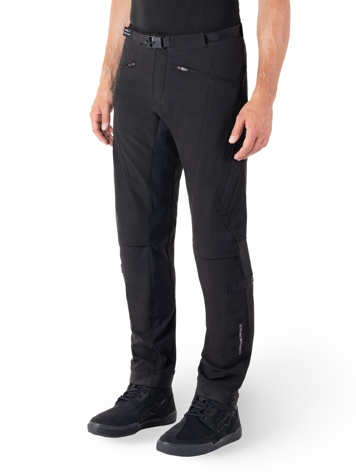 Alpinestars Alden Hose, Urbane Motorradhose, Schwarz, technisches Softshell-Gewebe mit normaler Passform, magnetische Gürtelschnalle, gesteppte Seiteneinsätze, Reißverschlusstaschen und dezentes Branding am Unterschenkel, getragen mit schwarzen Fahrerschuhen