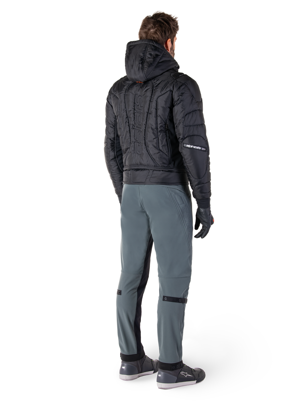 Alpinestars Alden Hose, urbane Motorradhose, Urban Gray, Rückansicht eines Fahrers in schwarzer gesteppter Kapuzenjacke und Motorrad-Sneakern, schmale Passform unterhalb des Knies mit schwarzen Stretcheinsätzen und Knieverstellern, modernes, von Streetwear inspiriertes Design.