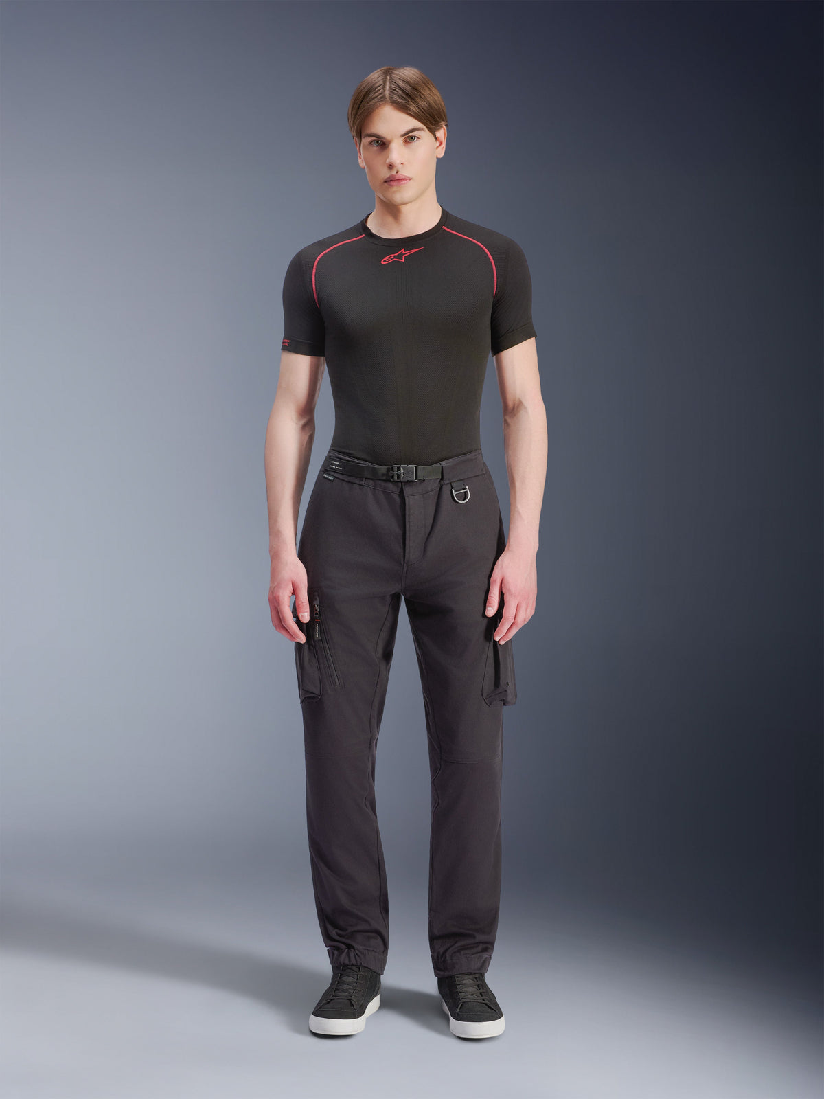 Alpinestars Flex-AST Cargo Broek - Short, zwart, gedragen door een mannelijk model met een zwart performance T-shirt en zwarte sneakers, stedelijke motorbroek met meerdere cargozakken, een taps toelopende pasvorm en een geïntegreerde riem met gesp en D-ring