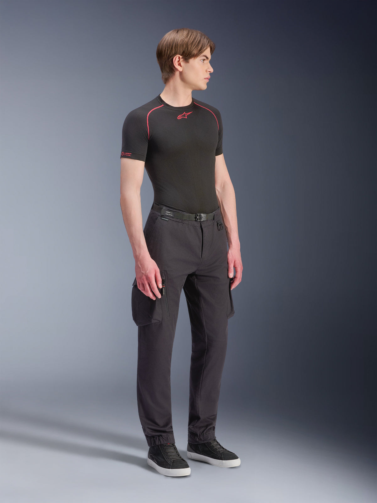 Mannelijk model draagt Alpinestars Flex-AST Cargo Pants - Short in het zwart, urban motorcargobroek met zijzakken en taps toelopende pijpen, gestyled met een zwarte base layer en sneakers, stedelijke motorkleding.