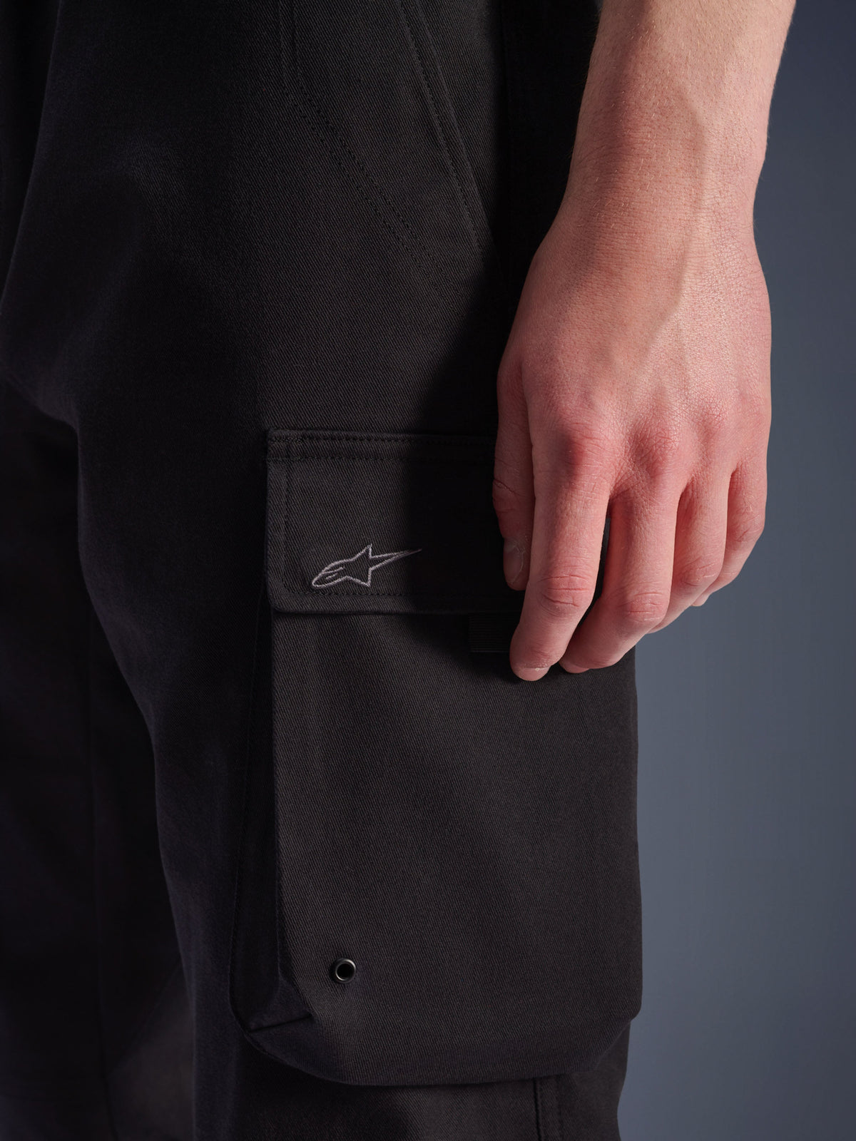 Alpinestars Flex-AST Cargo broek, urban motorbroek, zwart, close-up van de cargozak op de zijkant met geborduurd Astar-logo, metalen oogje en duurzame stretch twill-textuur