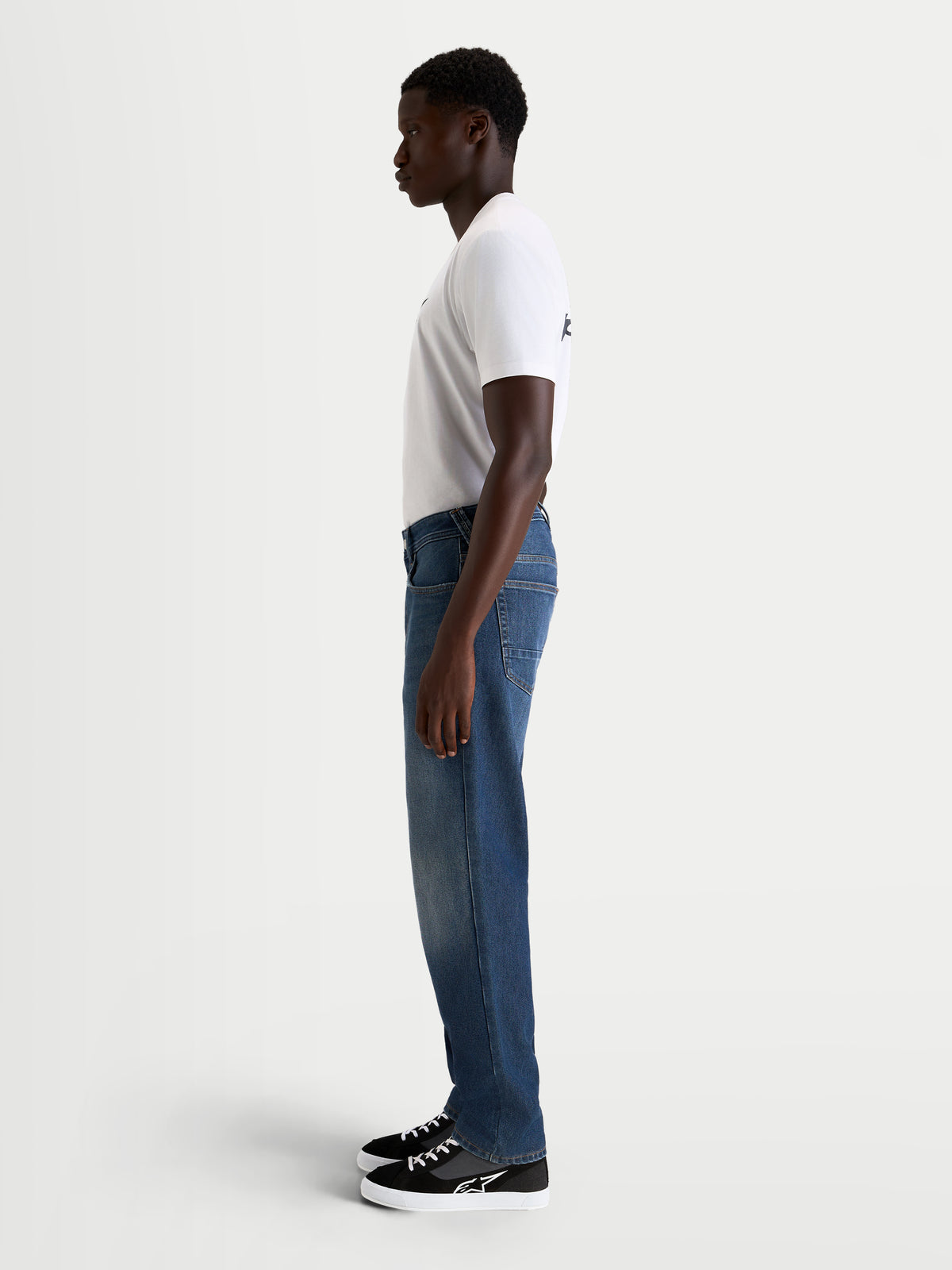 Alpinestars Flex Ast 8 Denim broek, urban motorjeans voor heren in middelblauw, zijaanzicht op model met wit t-shirt en casual motorlaarzen, AA-geclassificeerd enkellaags denim met stretchconstructie
