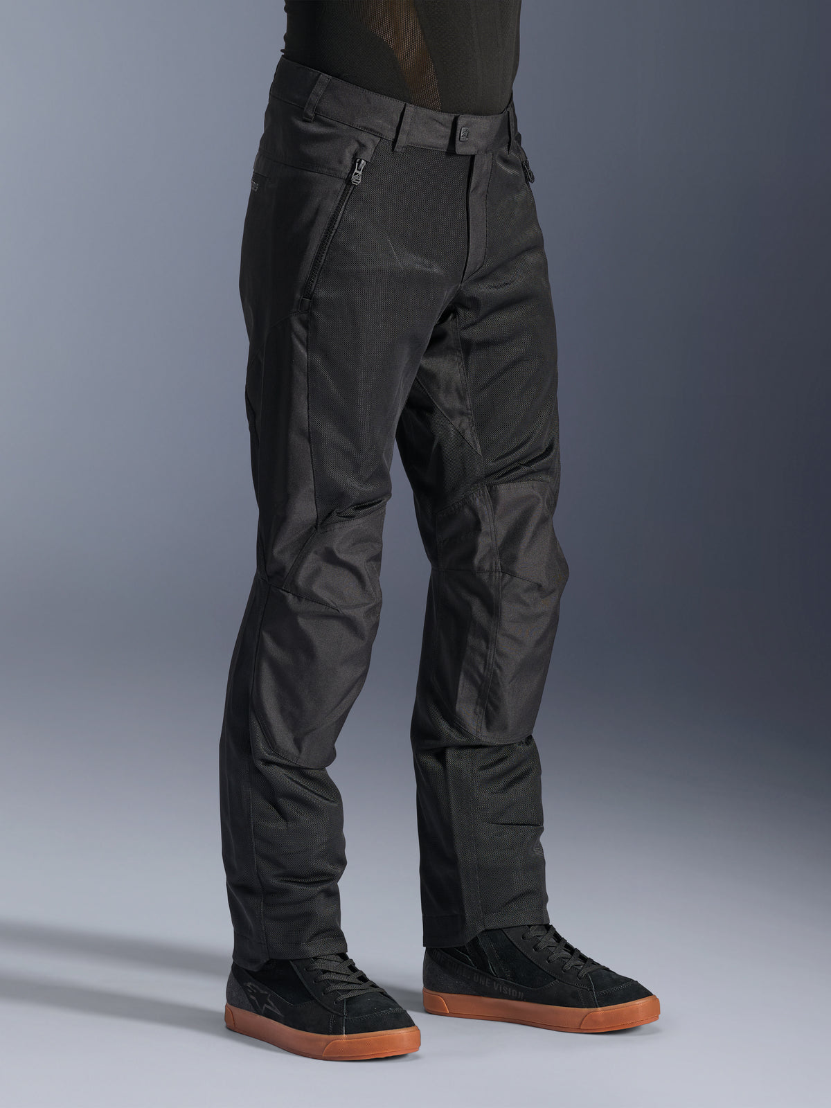Pantalon Alpinestars C-1 Air, pantalon de moto urbain, noir, doté de panneaux en maille technique respirante et d'une construction en polyfibre, porté avec des baskets noires, design épuré et fonctionnel pour la conduite en ville.