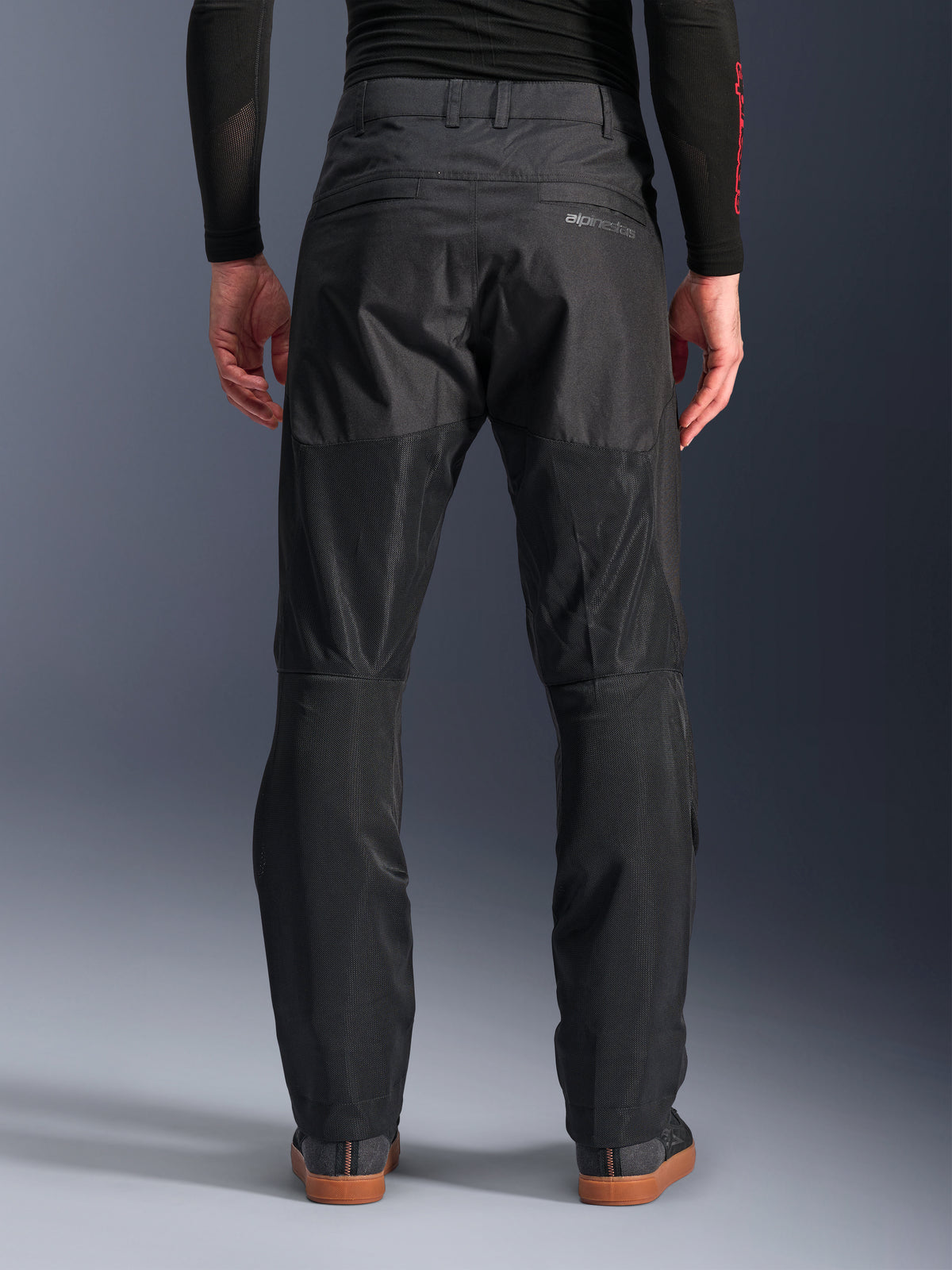 Alpinestars C-1 Air Pants, vue arrière, noir, pantalon de moto urbain en textile doté de larges panneaux en mesh technique à l'arrière des jambes pour la ventilation, design épuré avec logo discret, porté avec une couche de base noire et des chaussures de moto décontractées.