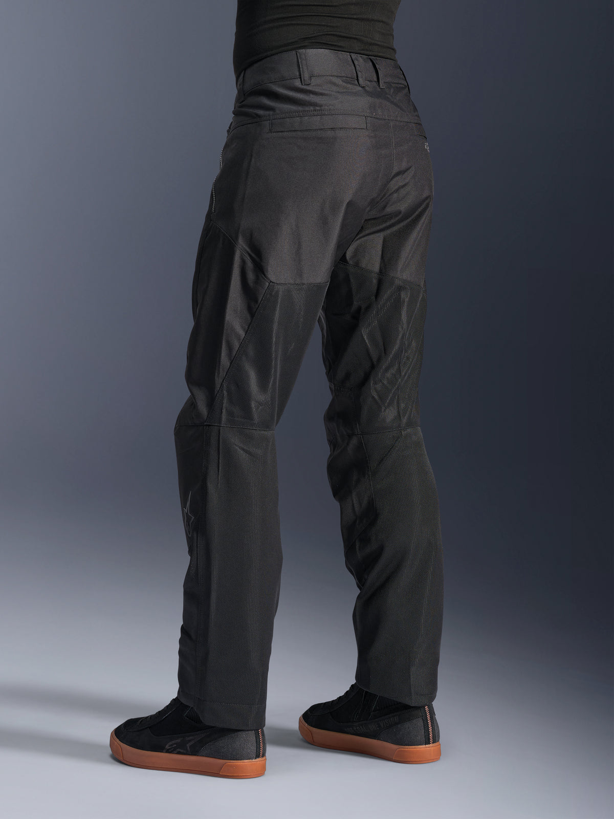 Pantalon Alpinestars C-1 Air, pantalon de moto urbain, noir, vue arrière montrant de larges panneaux en mesh technique à l'arrière des jambes pour la ventilation, style épuré pour la conduite urbaine, porté avec des baskets noires décontractées.