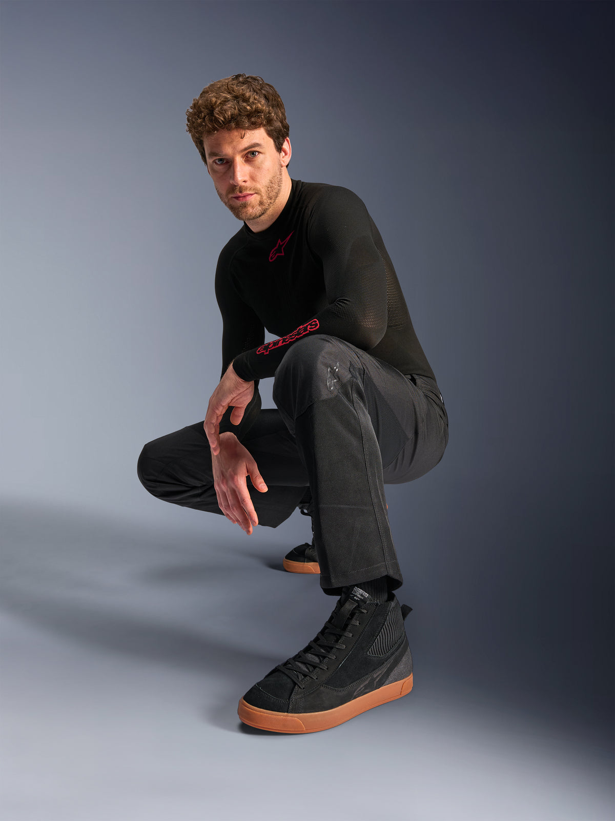 Alpinestars C-1 Air Pants, pantalon de moto urbain, noir, modèle accroupi dans un studio, porté avec un haut noir à manches longues et des baskets noires à semelles marron, doté de panneaux en mesh respirant et d'un style urbain contemporain