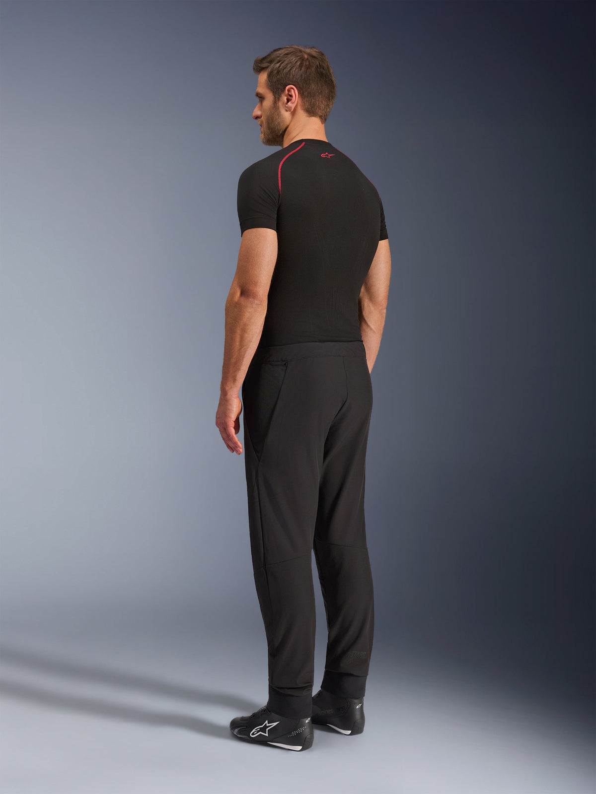 Alpinestars Aeroshell Airflow Hose, schwarze Motorradhose, leichtes Softshell-Textil, kombiniert mit einem schwarzen technischen Kurzarmshirt und schwarzen Motorradstiefeln, seitliche Rückansicht zeigt die schmale Passform und ergonomische Konstruktion.
