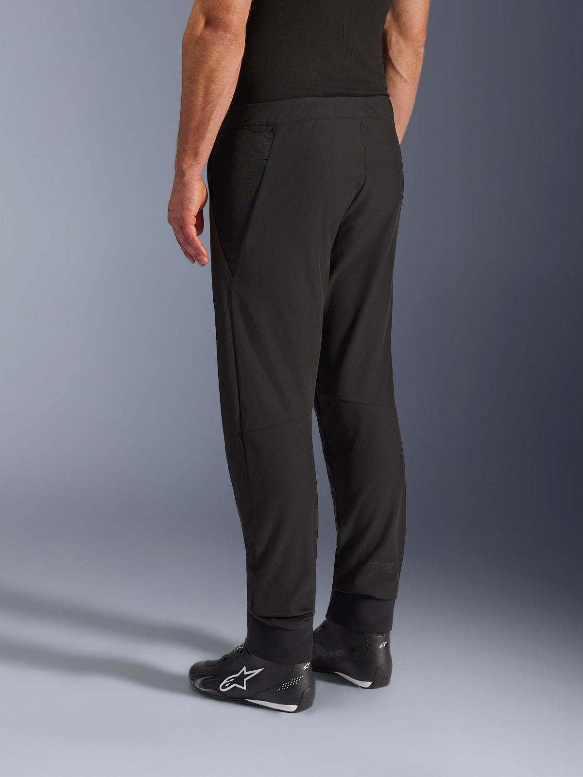 Alpinestars Aeroshell Airflow Hose, schwarze Motorradhose, Dreiviertel-Rückansicht, leichte Stretch-Softshell-Konstruktion mit integrierten Mesh-Einsätzen, verstellbare Knöchelbündchen, getragen mit schwarzen Alpinestars Motorradschuhen