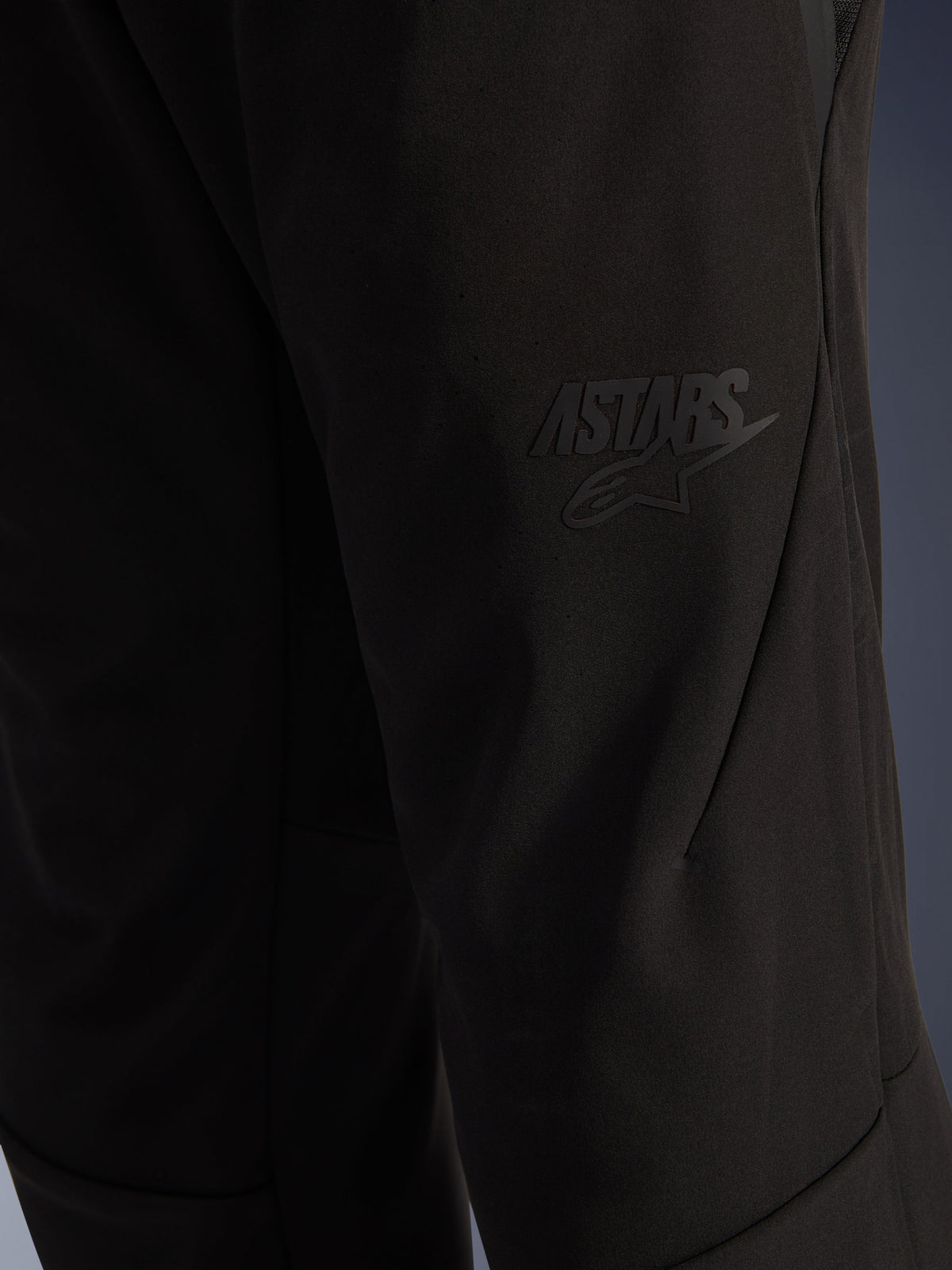 Alpinestars Aeroshell Airflow Hose, schwarze Motorradhose, Nahaufnahme des Oberschenkelbereichs mit erhabenem schwarzem ASTARS-Logo, leichtes Stretch-Softshell-Material mit ergonomischem Nahtdesign