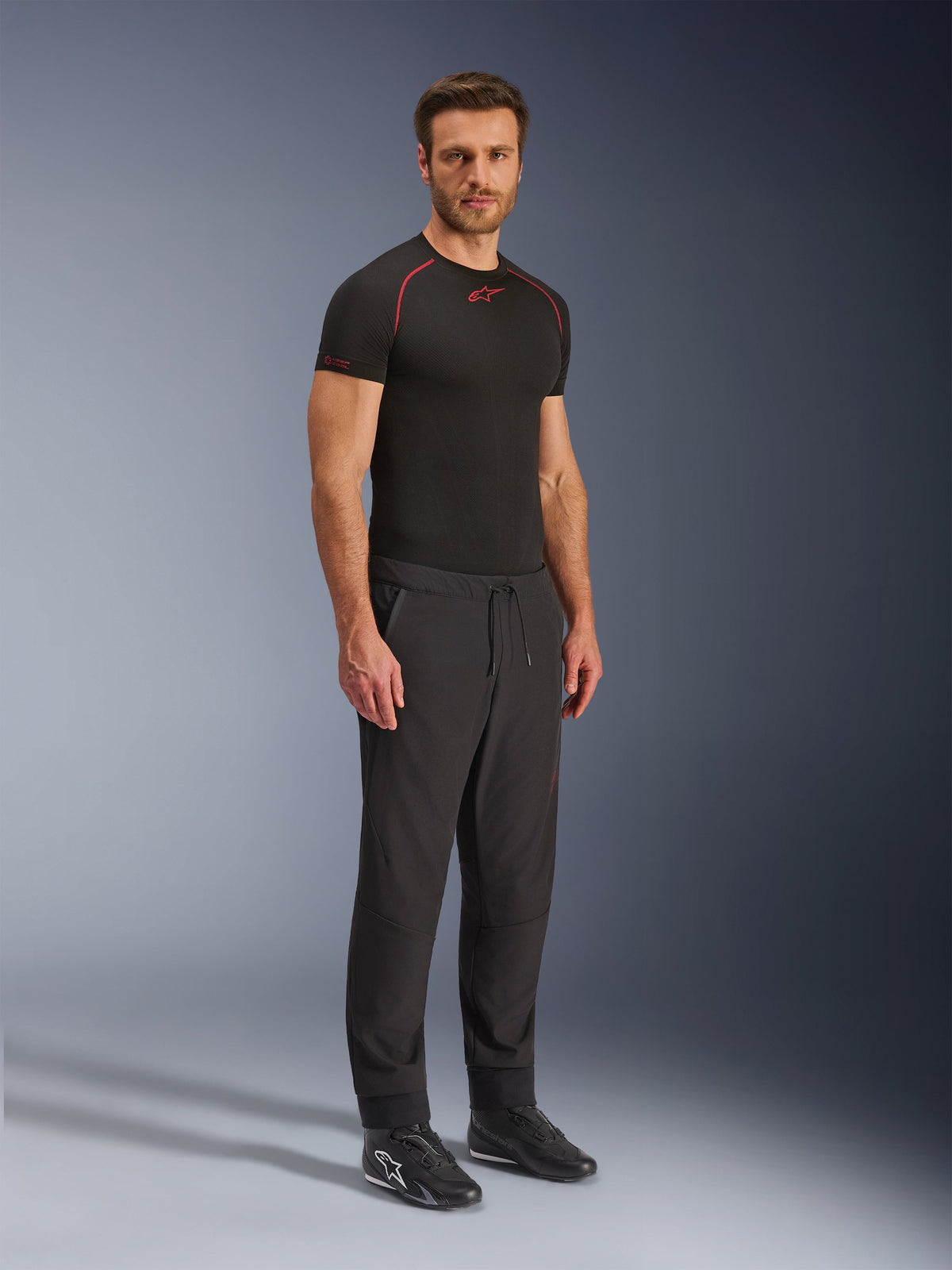 Alpinestars Aeroshell Airflow Hose, schwarze Motorradhose, Ganzkörper-Studioaufnahme eines Models in Softshell-Hose mit Kordelzug am Bund und elastischen Bündchen, kombiniert mit einem schwarzen Alpinestars Baselayer-Shirt und schwarzen Motorradschuhen.