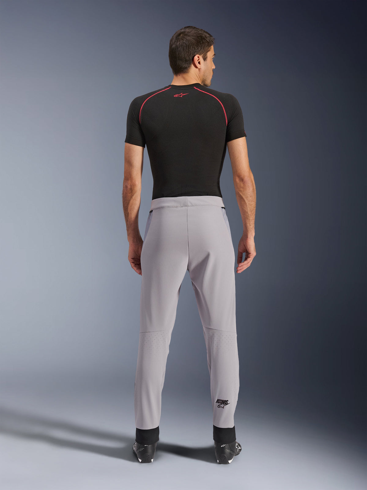 Pantalon Alpinestars Aeroshell Airflow, Gris moyen, vue arrière montrant les zones perforées au laser sur les mollets, construction en softshell extensible léger, porté avec un t-shirt noir à manches courtes et des bottes de moto, coupe sportive pour la conduite par temps chaud