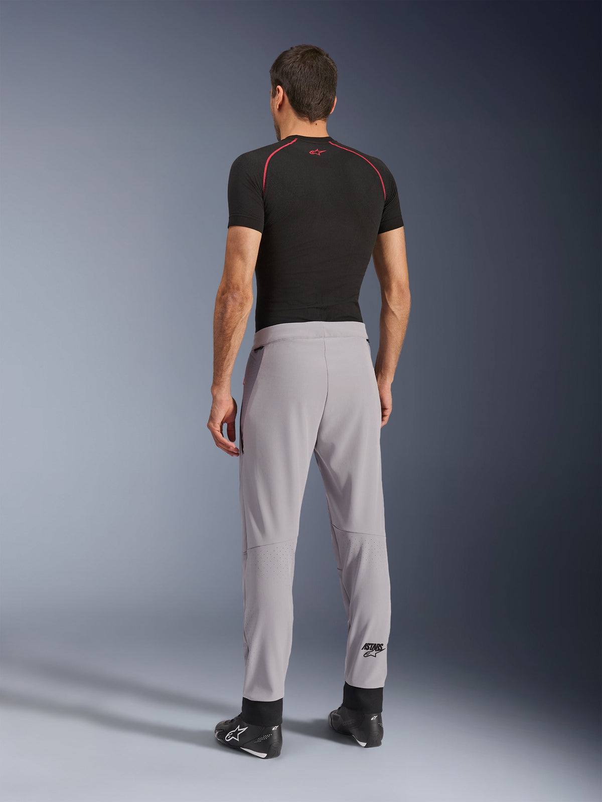 Vue arrière d'un mannequin portant le pantalon Aeroshell Airflow d'Alpinestars en Mid Gray, associé à un haut de couche de base noir et des chaussures noires, mettant en évidence les zones perforées au laser sur les mollets et la coupe sportive athlétique.