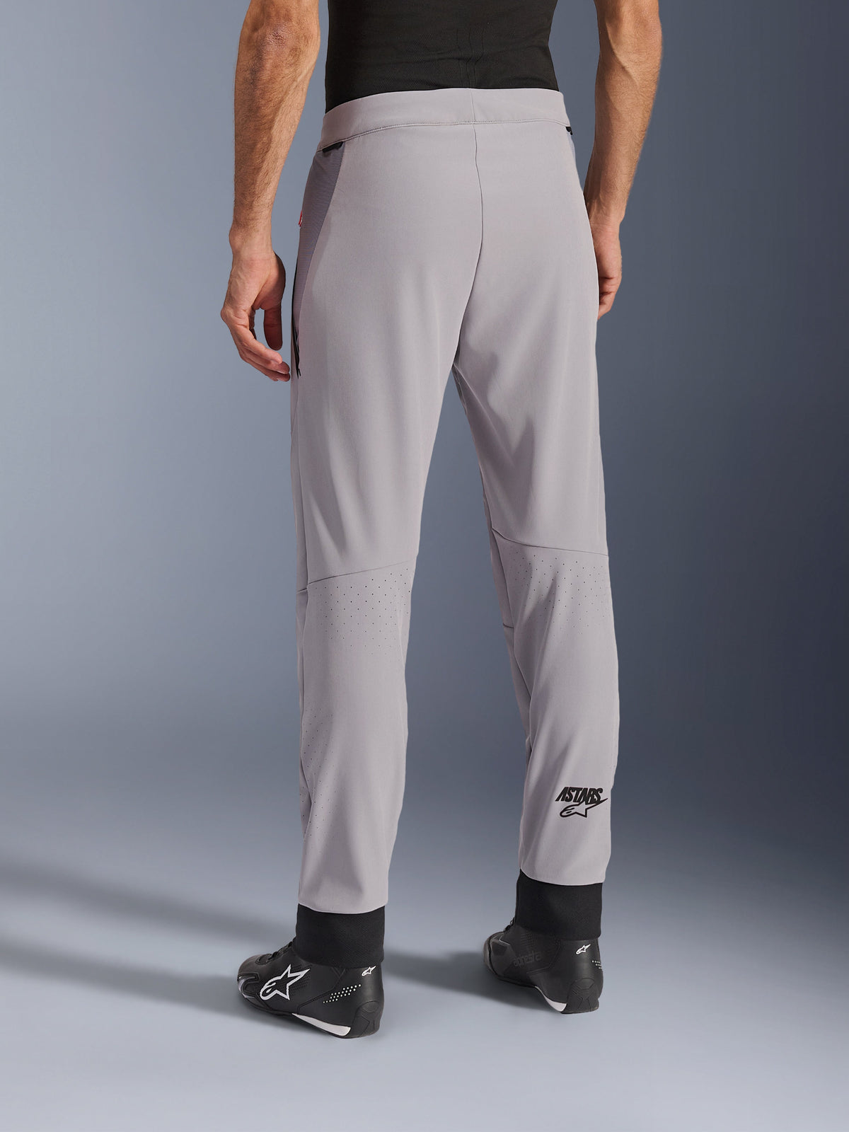 Pantalon Alpinestars Aeroshell Airflow, Gris Moyen, vue arrière montrant les zones perforées au laser sur les mollets, logo ASTARS sur le bas de la jambe, poignets de cheville noirs réglables, porté avec des bottes de moto noires, pantalon textile léger pour la conduite sportive